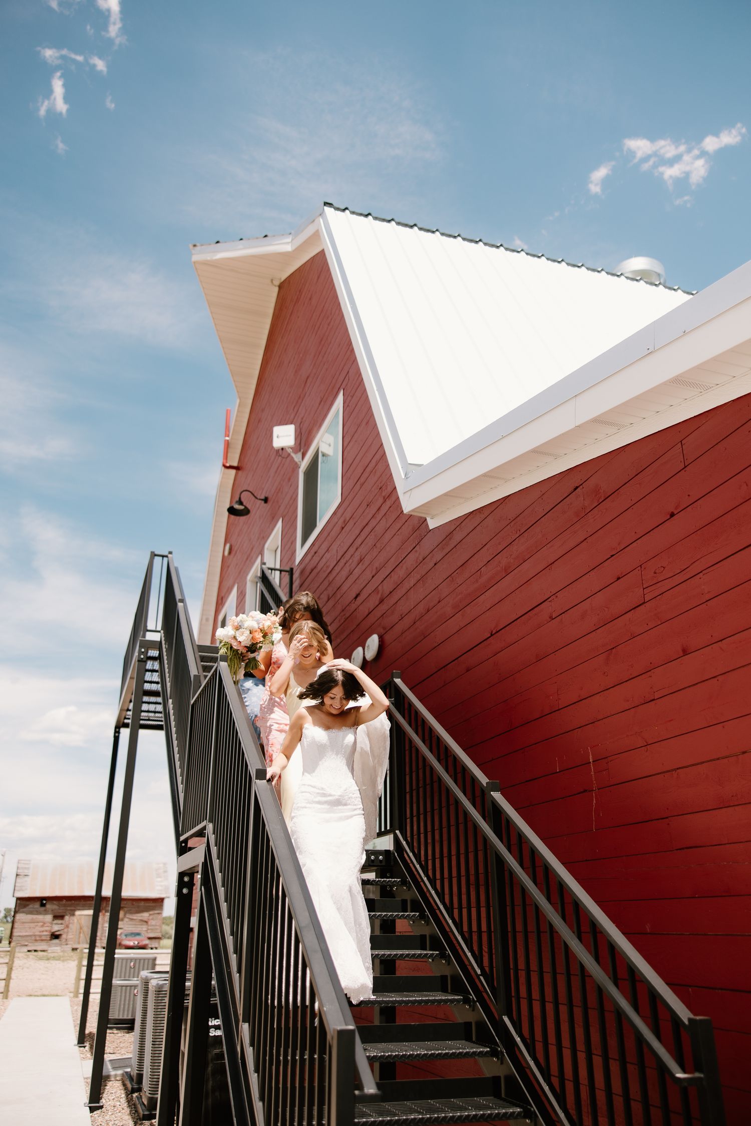 Two brides descend metal stairs outside a red barn venue under a bright blue sky, one in a white gown holding a bouquet.