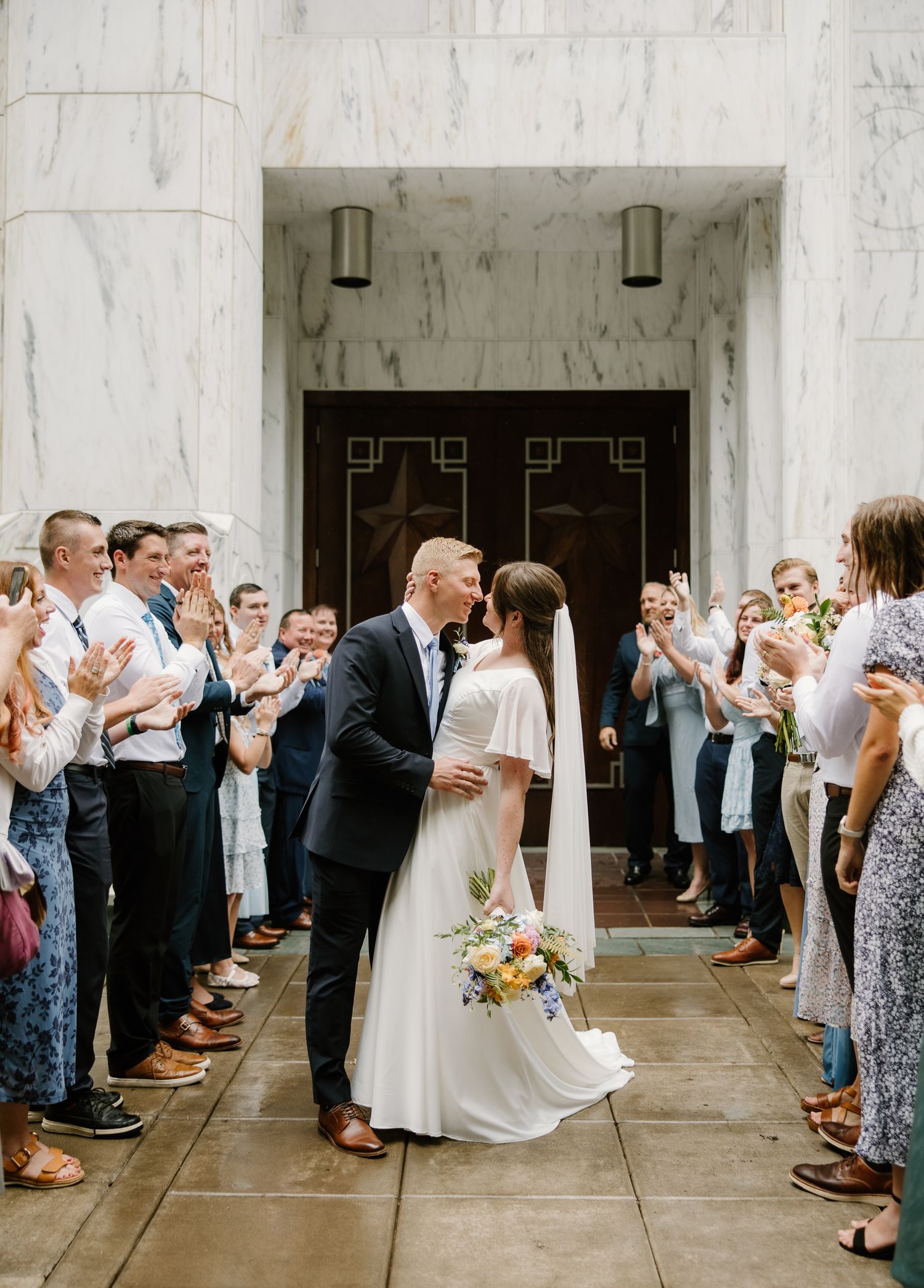 Bride and groom share a kiss while guests applaud in a grand marble building exit.