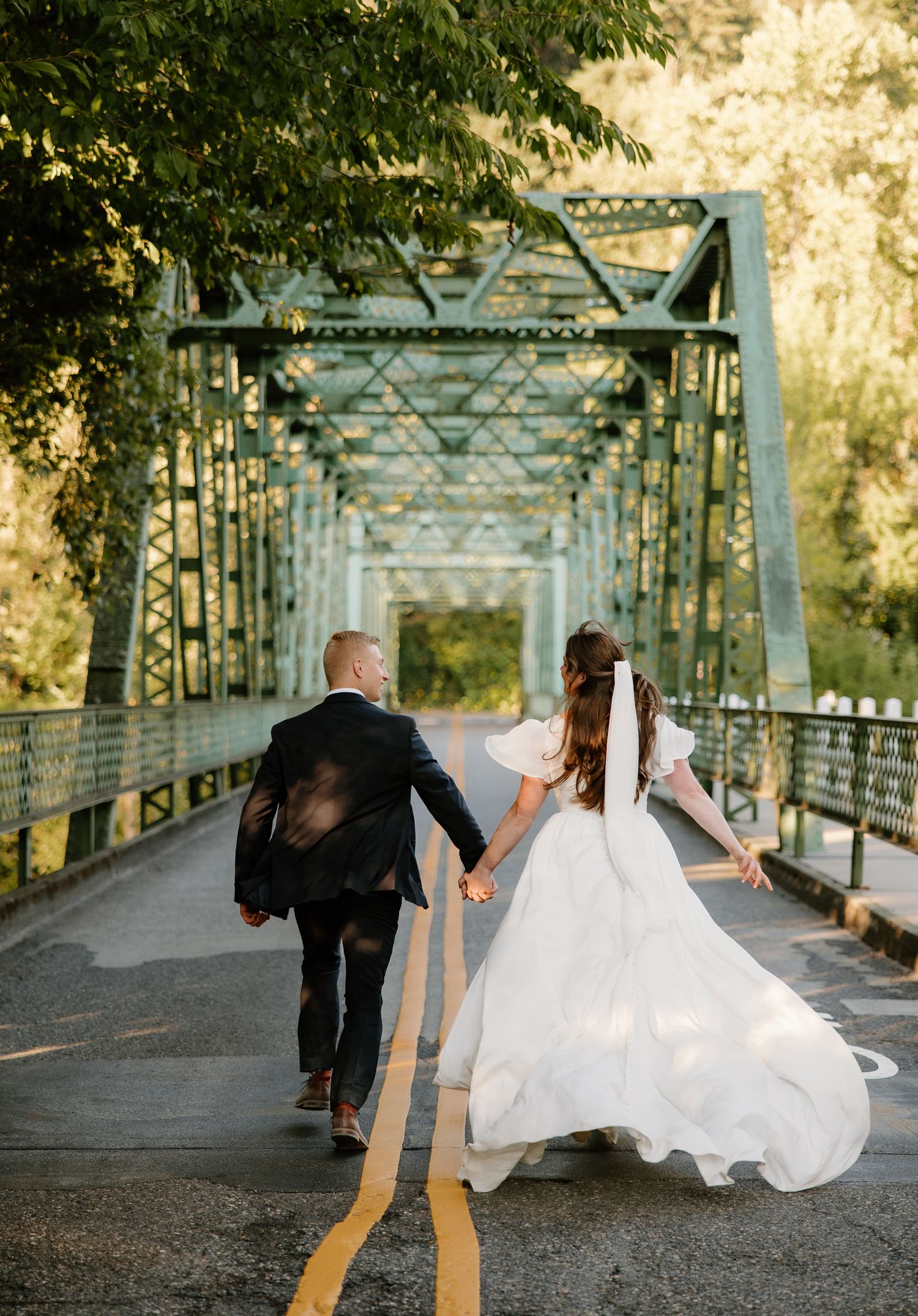 Bride and groom holding hands walking across vintage green steel bridge on their wedding day.