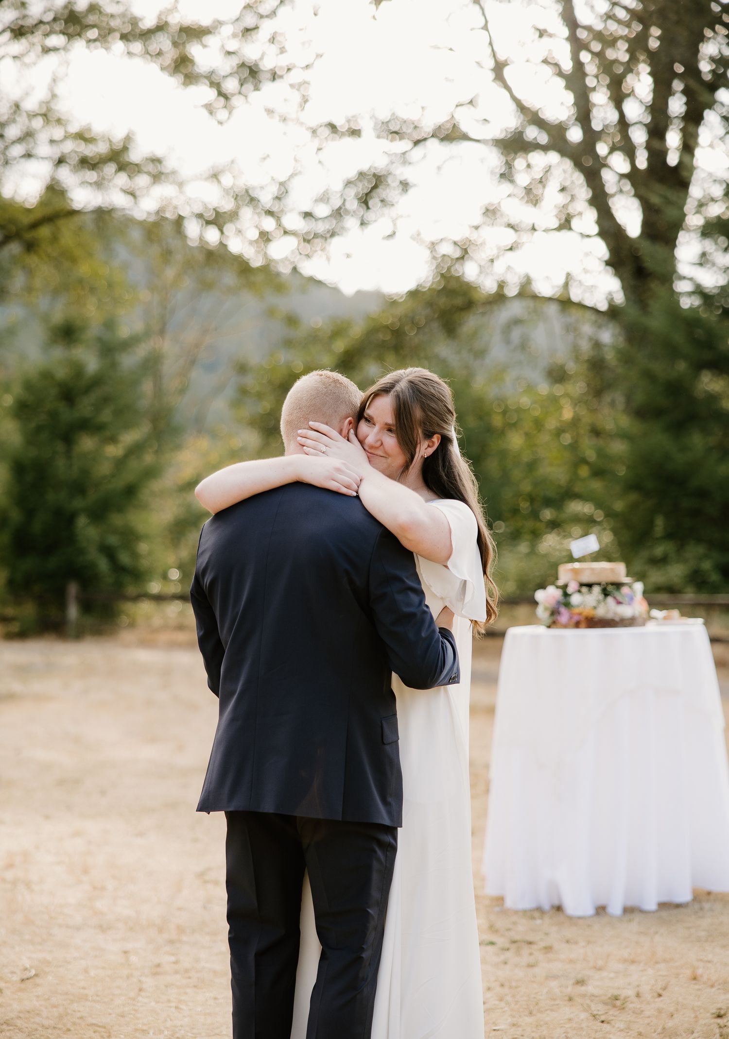 Bride and groom share an emotional embrace during their outdoor wedding reception near tall trees.