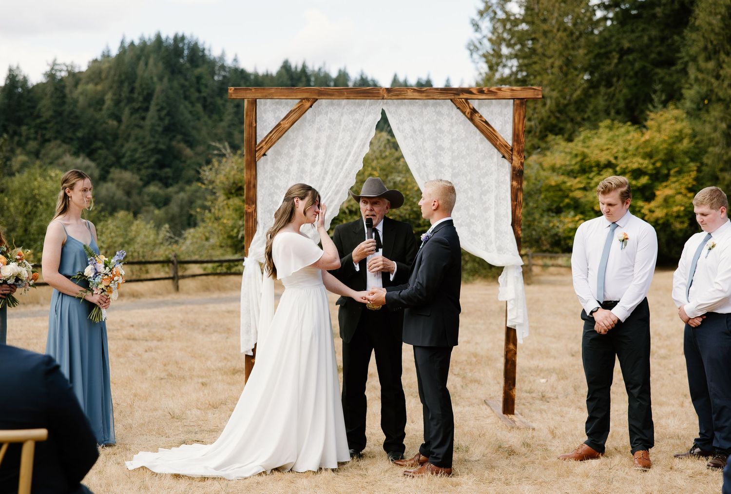 Bride and groom exchange vows under rustic wooden arch in scenic outdoor meadow ceremony.