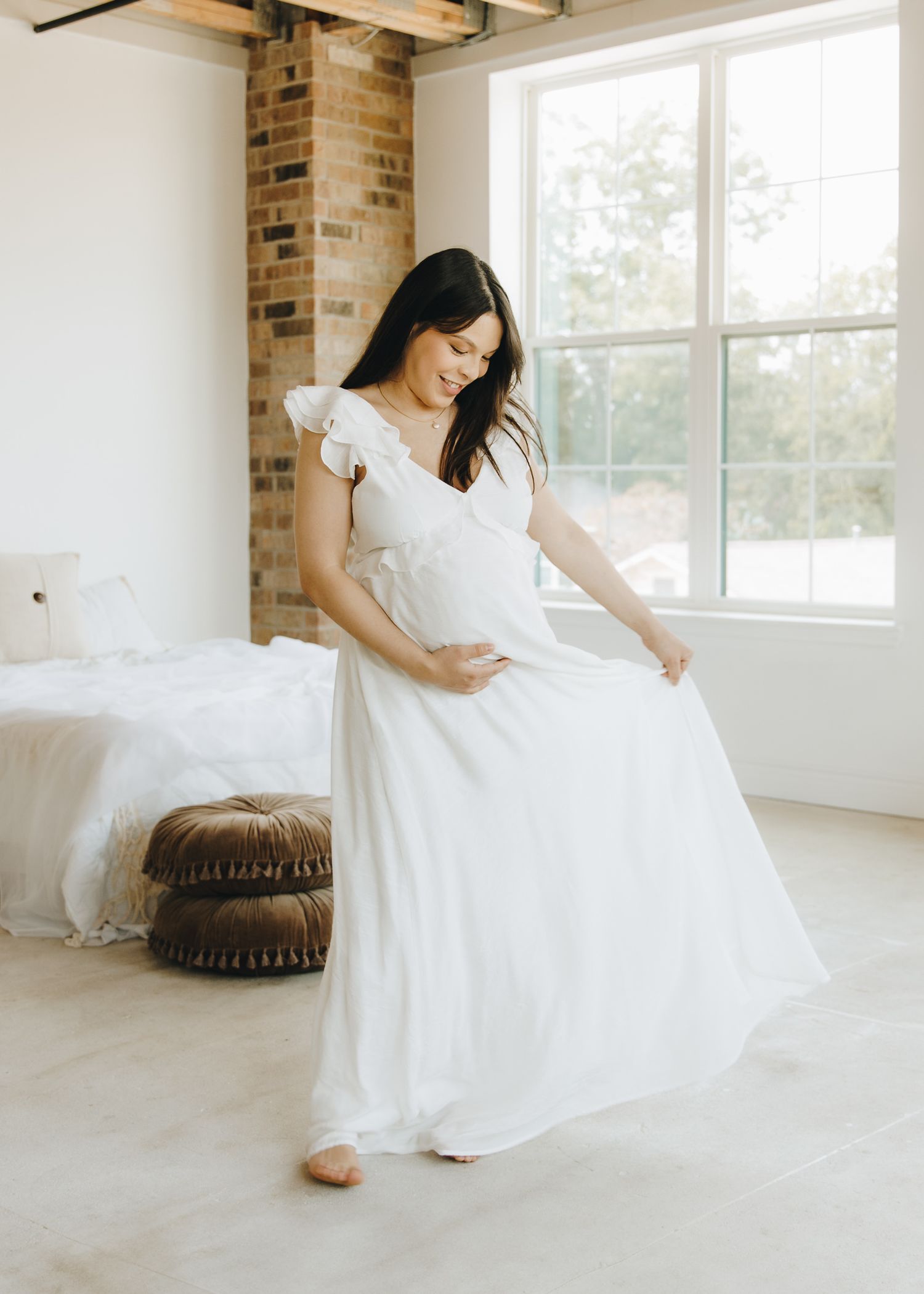 Smiling pregnant woman in flowing white dress twirling in a bright, airy loft studio.