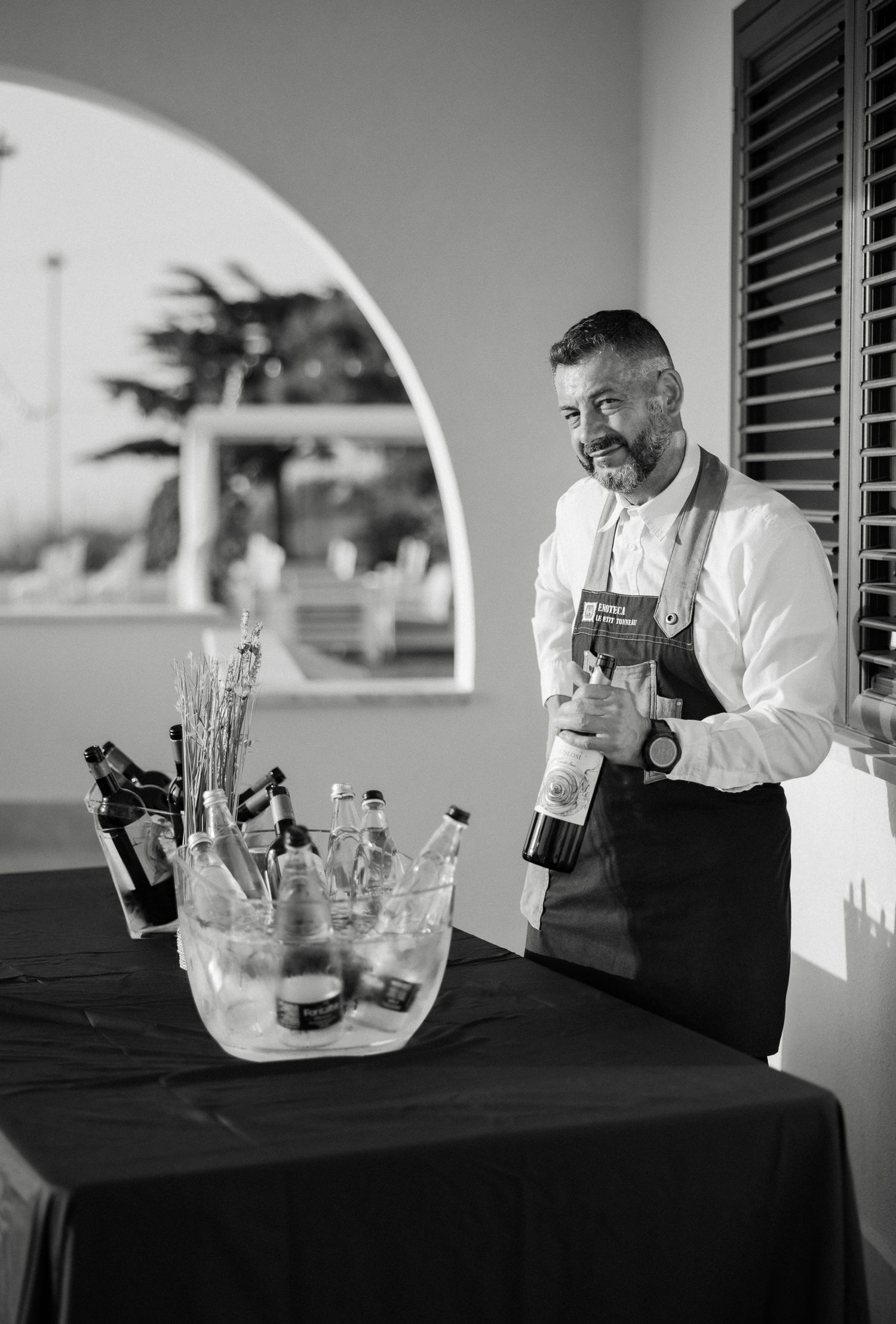 Black and white photo of a bartender holding a wine bottle at an outdoor wedding bar.