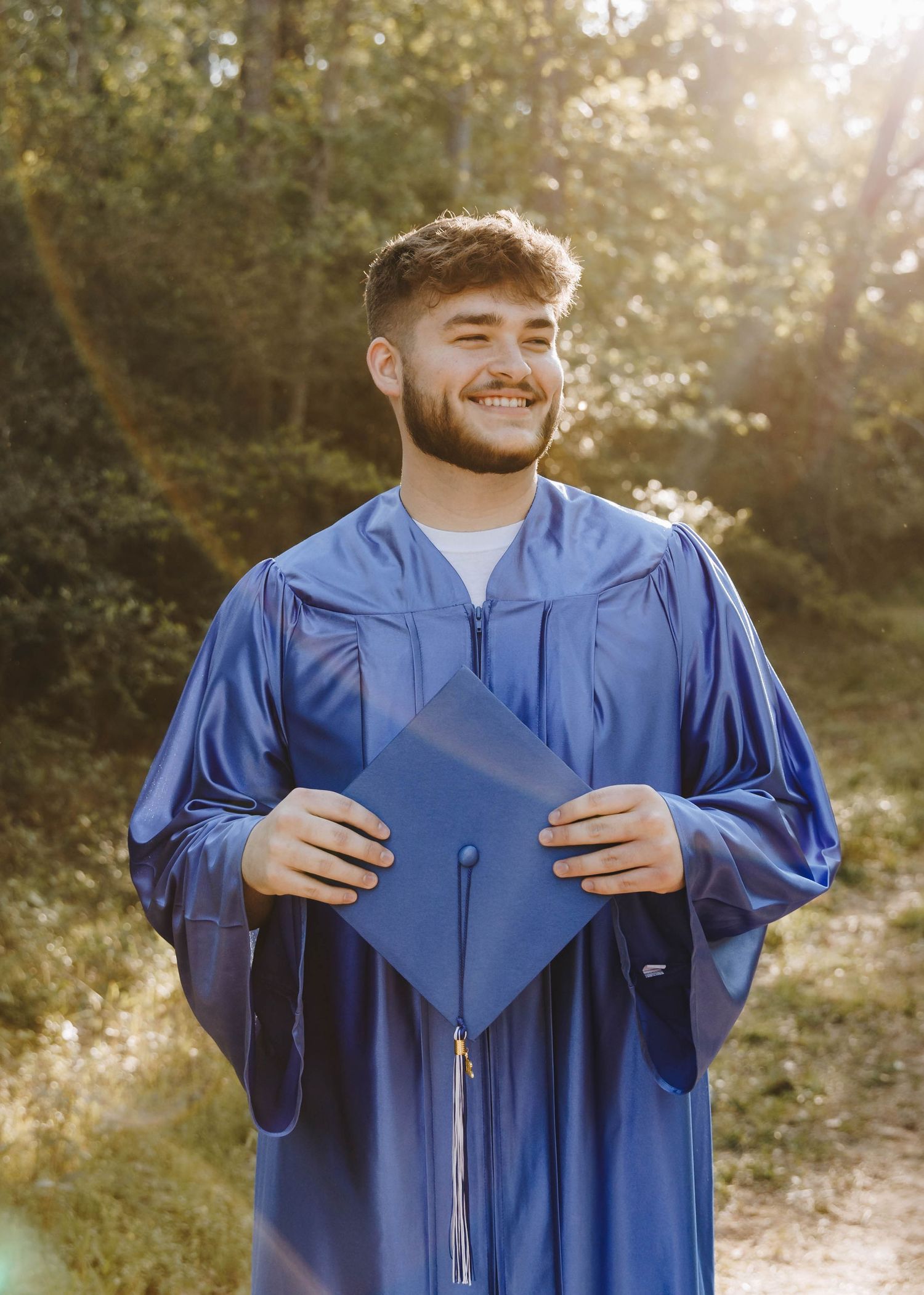 Happy young man in blue graduation cap and gown holding his diploma cover in sunny woods.