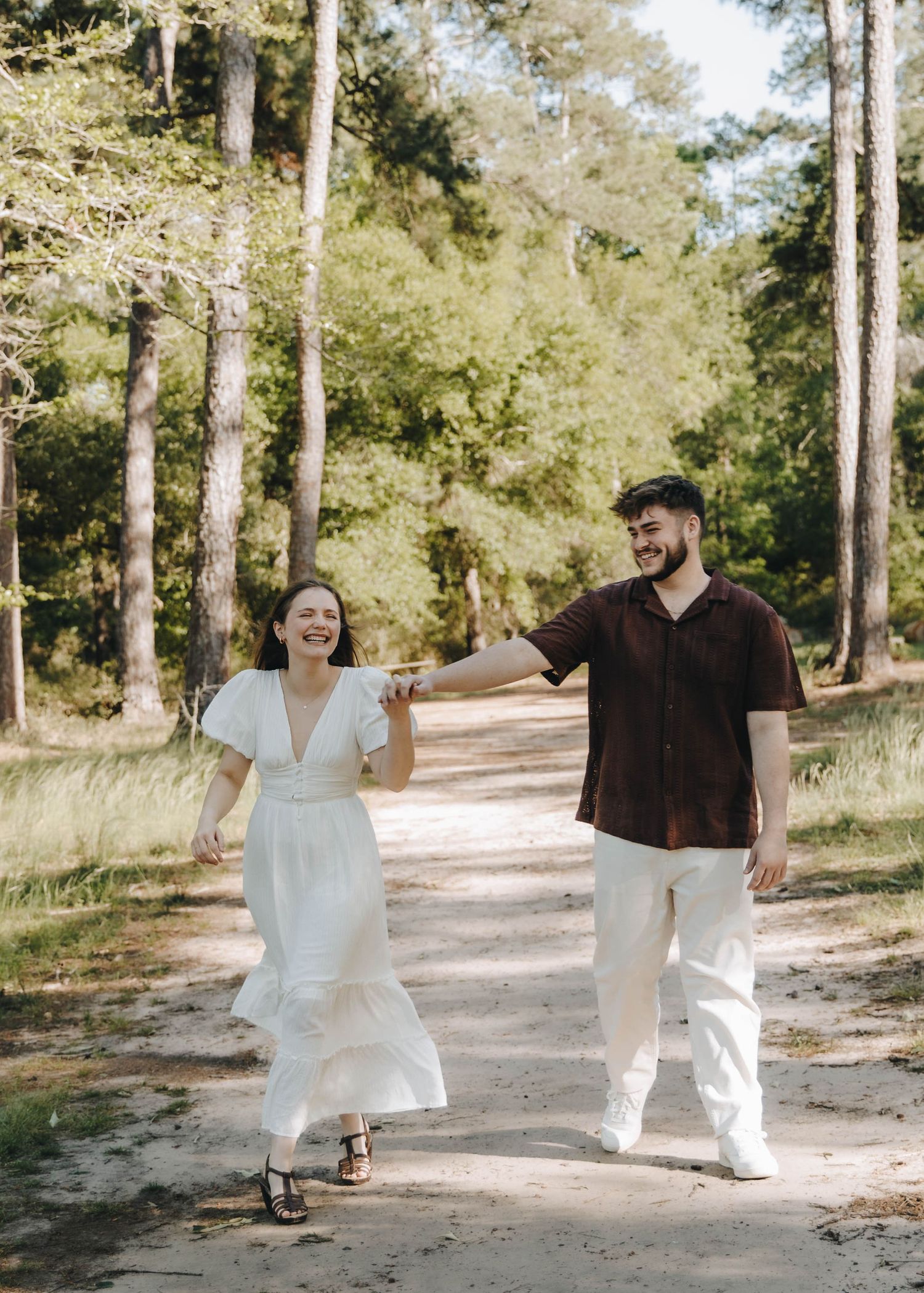 Joyful couple holding hands walking down a sunlit forest path, woman in white dress.