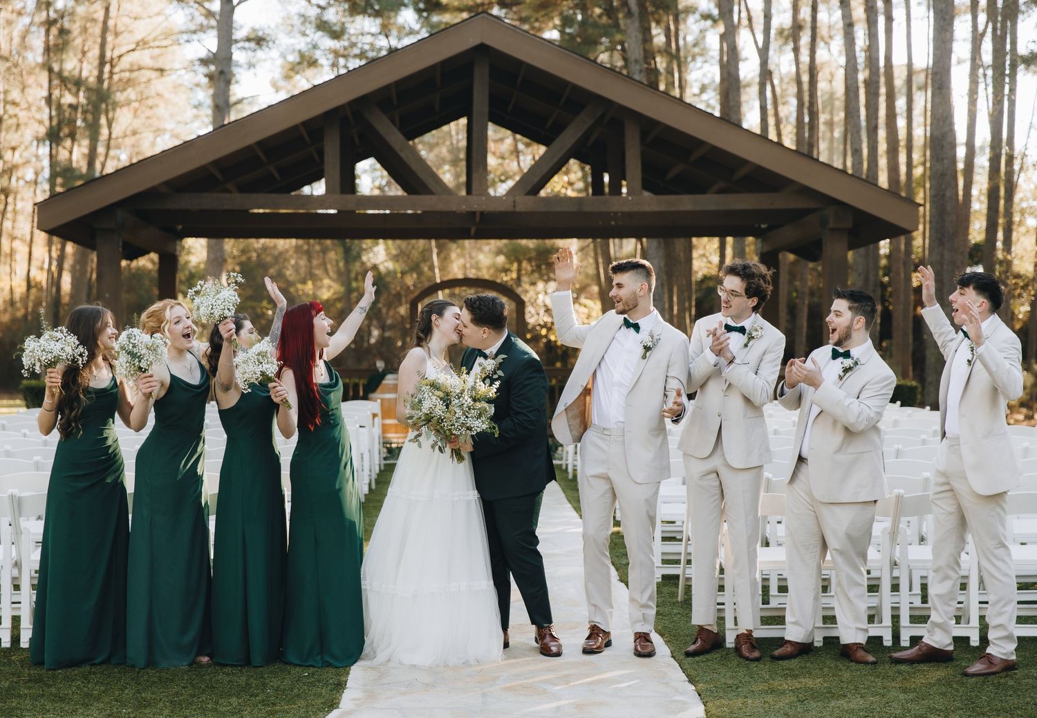 Bride and groom share a kiss at outdoor wedding ceremony under wooden pavilion with bridal party cheering.