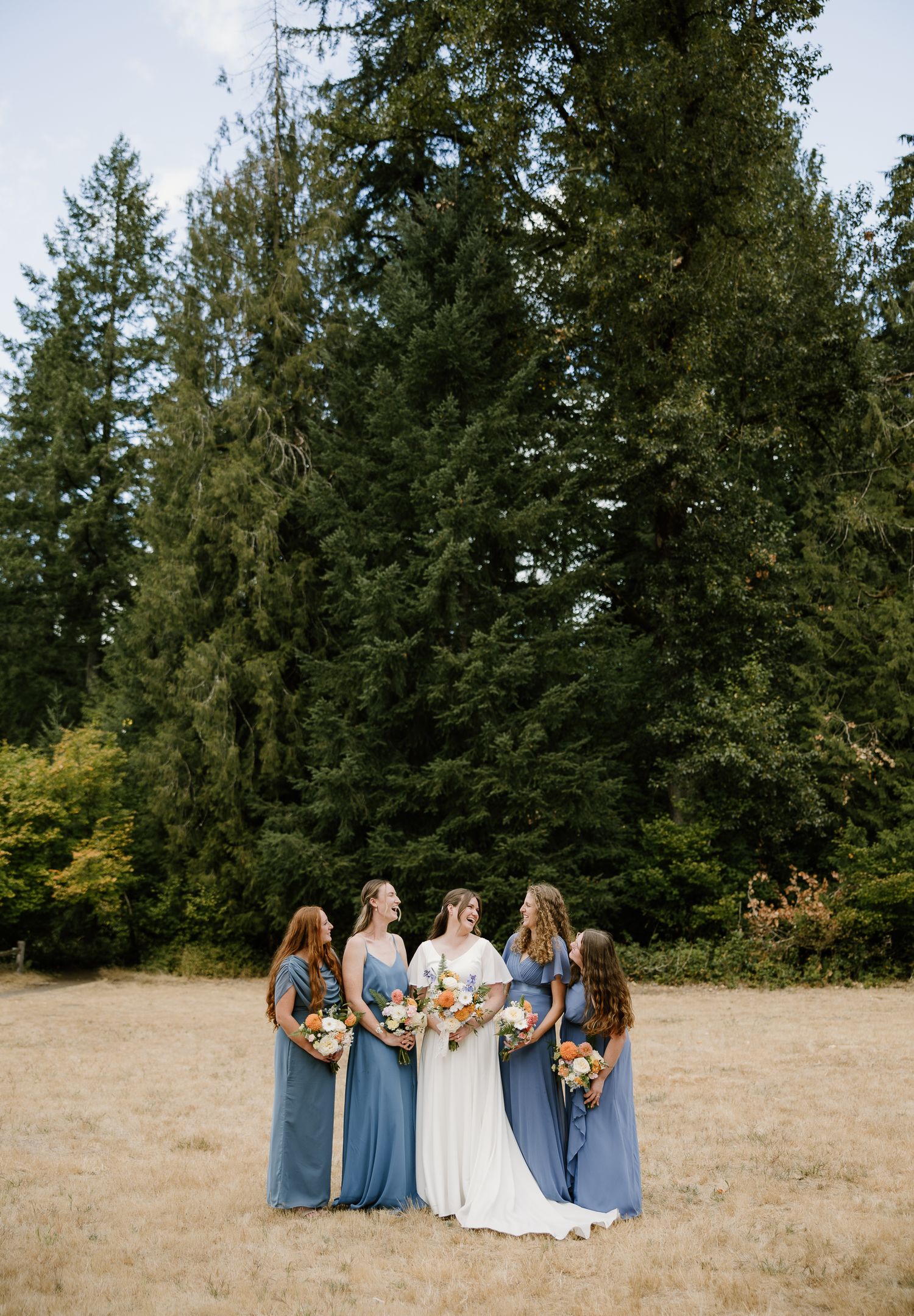 Bride poses with four bridesmaids in blue gowns holding colorful bouquets in front of tall evergreen trees.