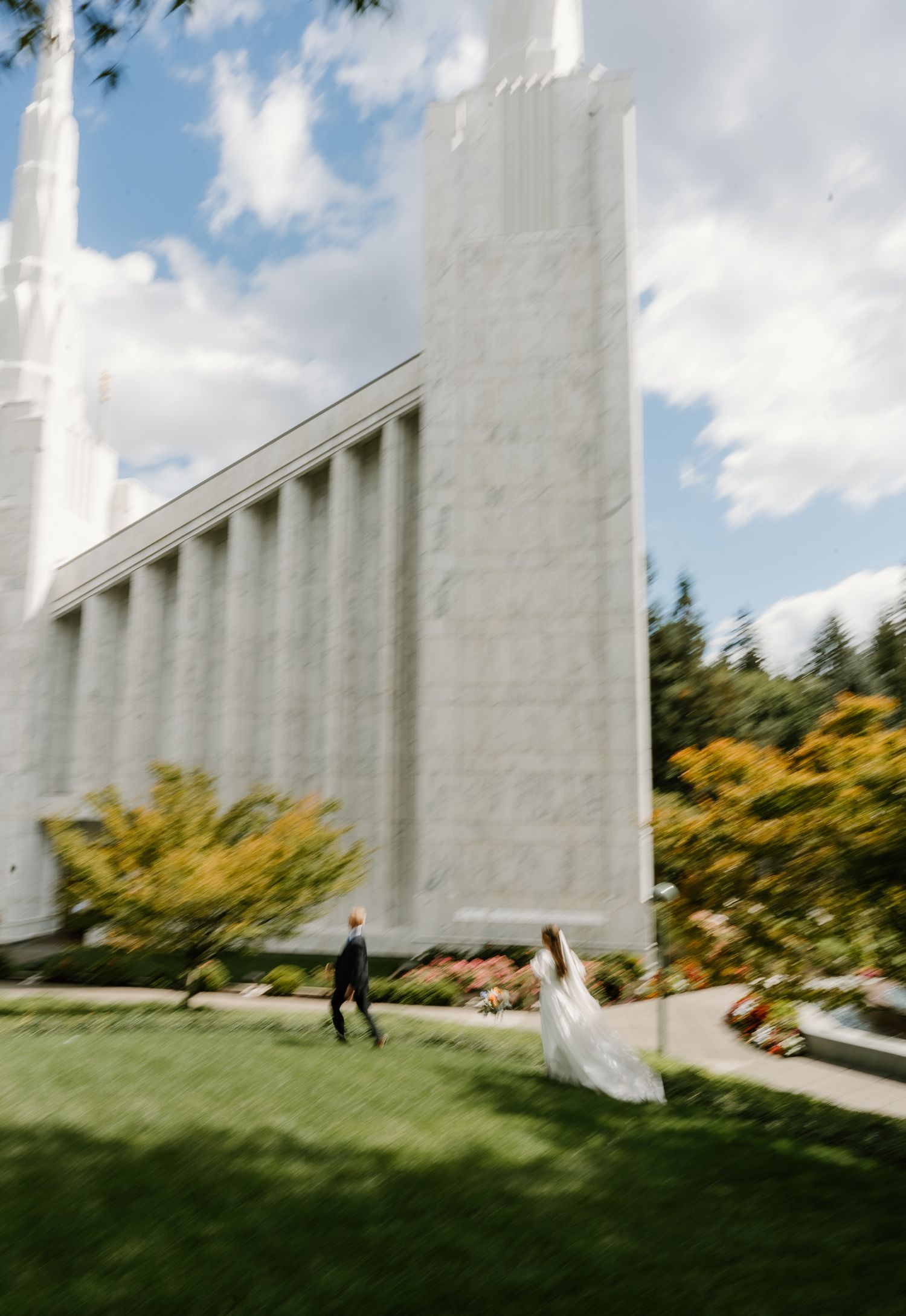 Bride in white gown and groom walk across green lawn beside tall white marble temple columns.