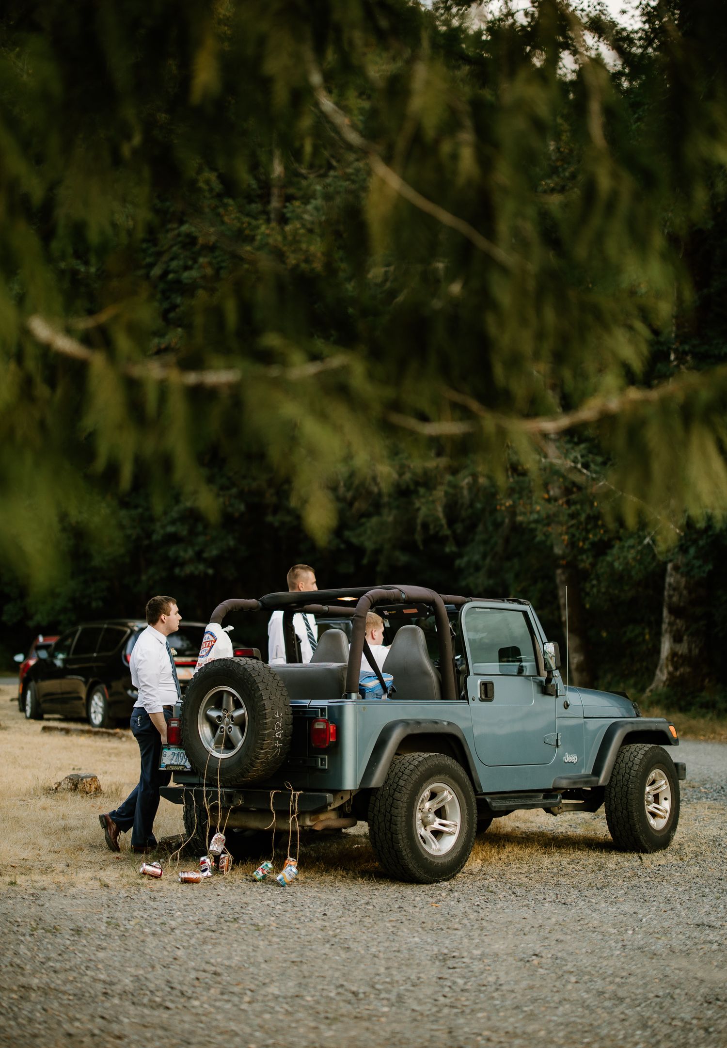 Two men stand beside a blue Jeep Wrangler decorated with wedding cans on a gravel path.