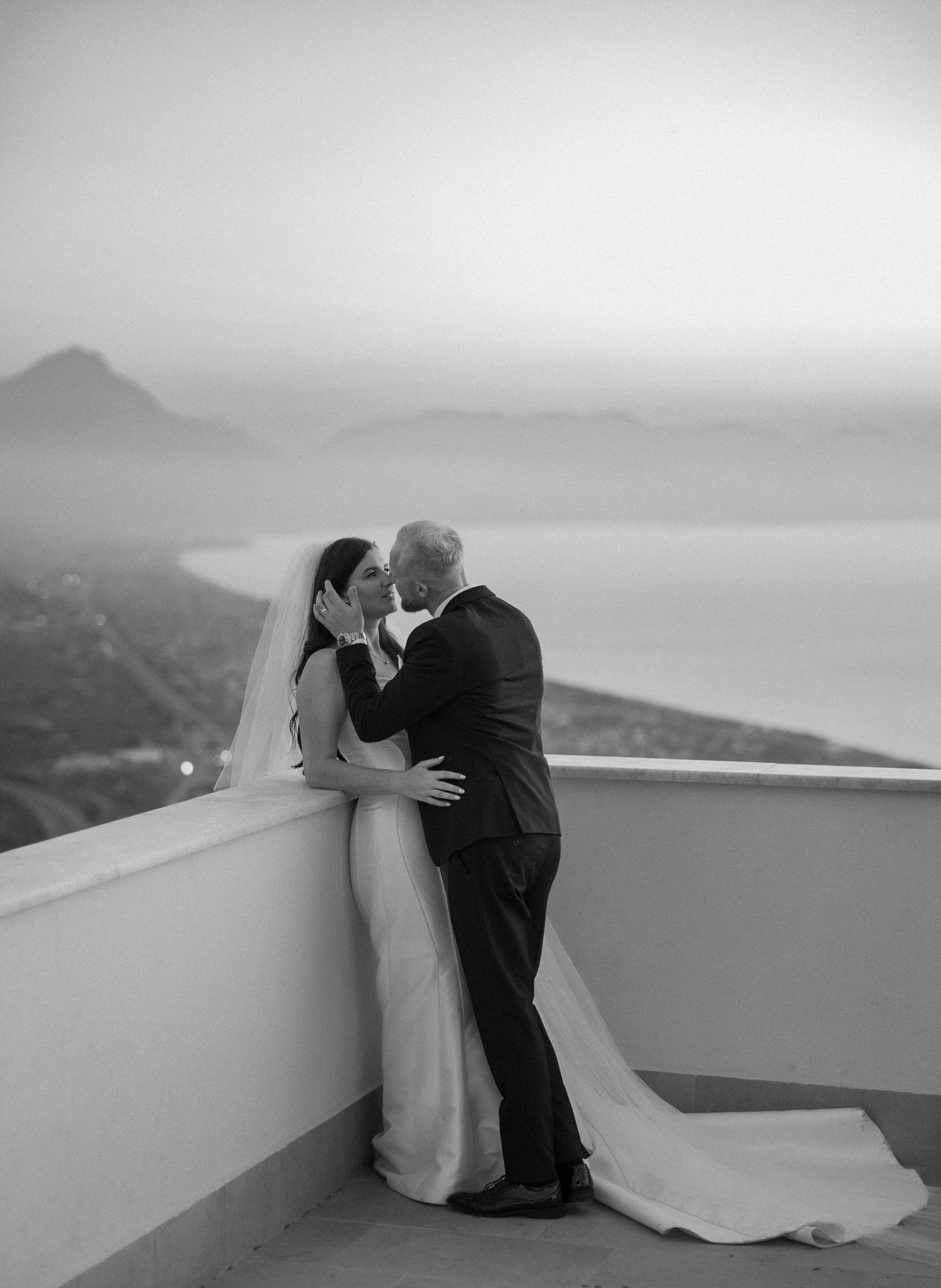 Black and white photo of bride and groom sharing a kiss on a terrace with misty coastal views.