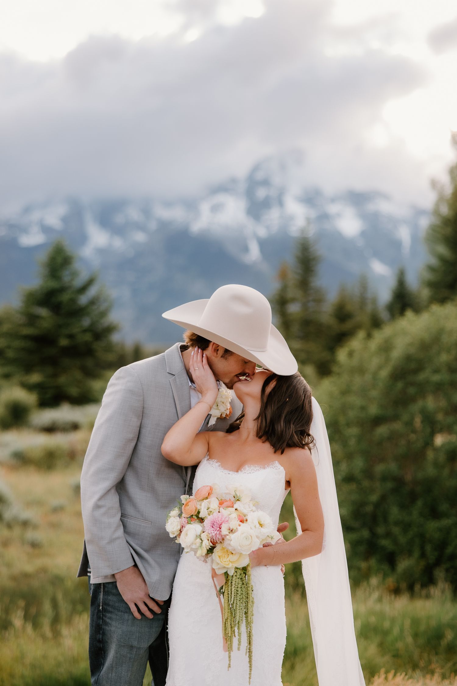 Bride and groom share a kiss with snow-capped mountain peaks and pine trees in the background.