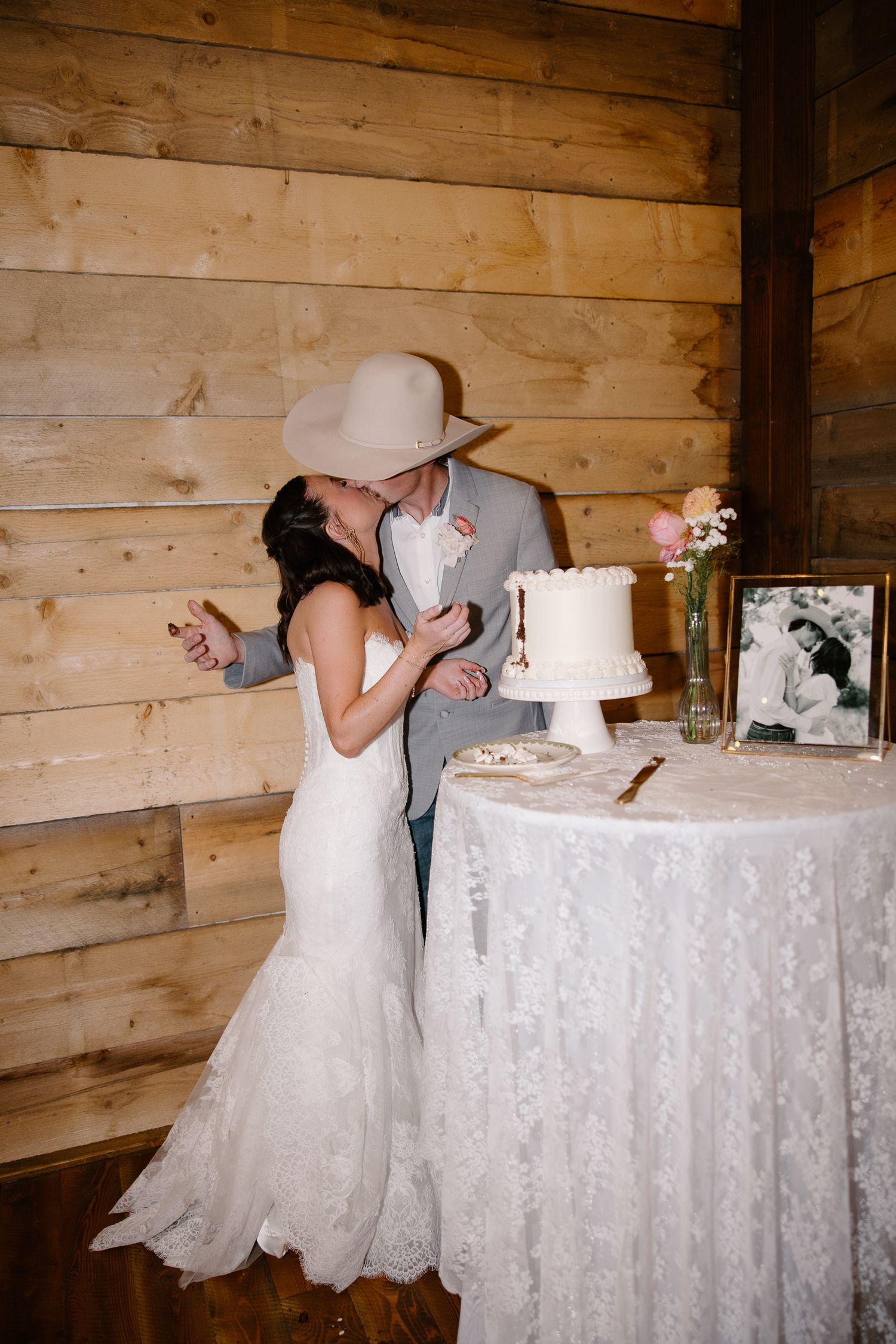 Bride and groom kiss while cutting white wedding cake at rustic barn reception.