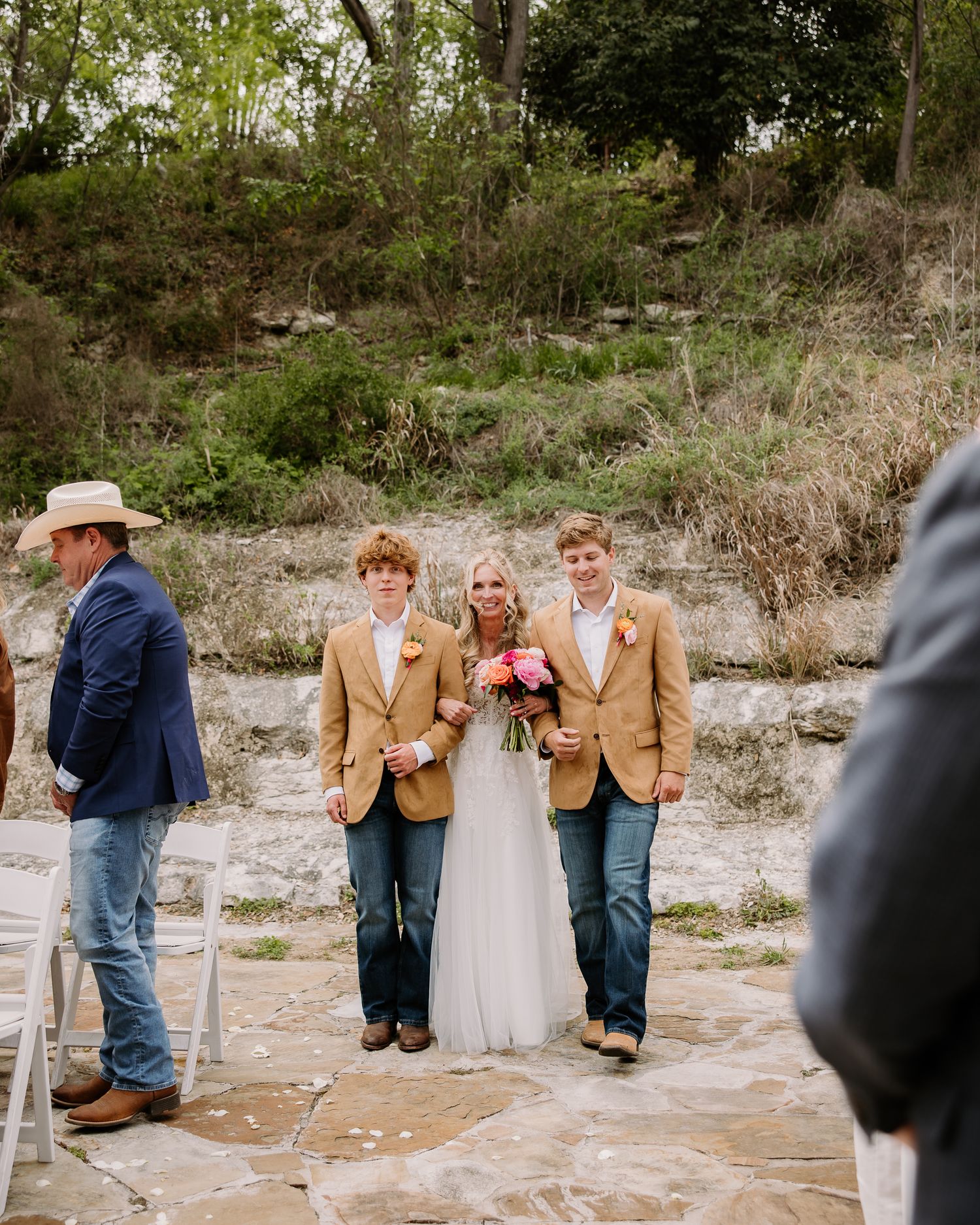 Bride walks down outdoor wedding aisle escorted by two young men in tan blazers and jeans, holding pink bouquet.