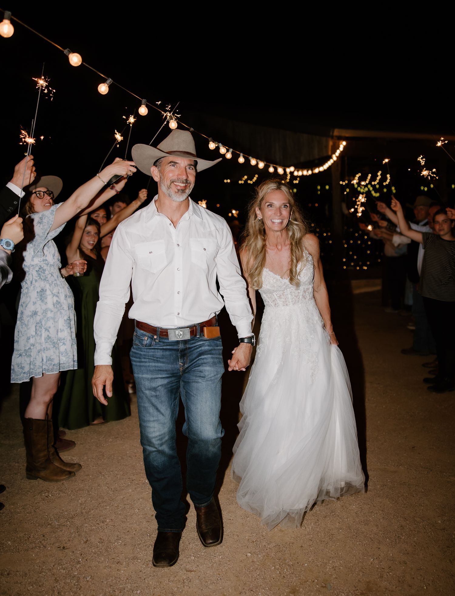 Smiling bride and groom in cowboy hat walk through sparkler send-off under string lights at outdoor western wedding.
