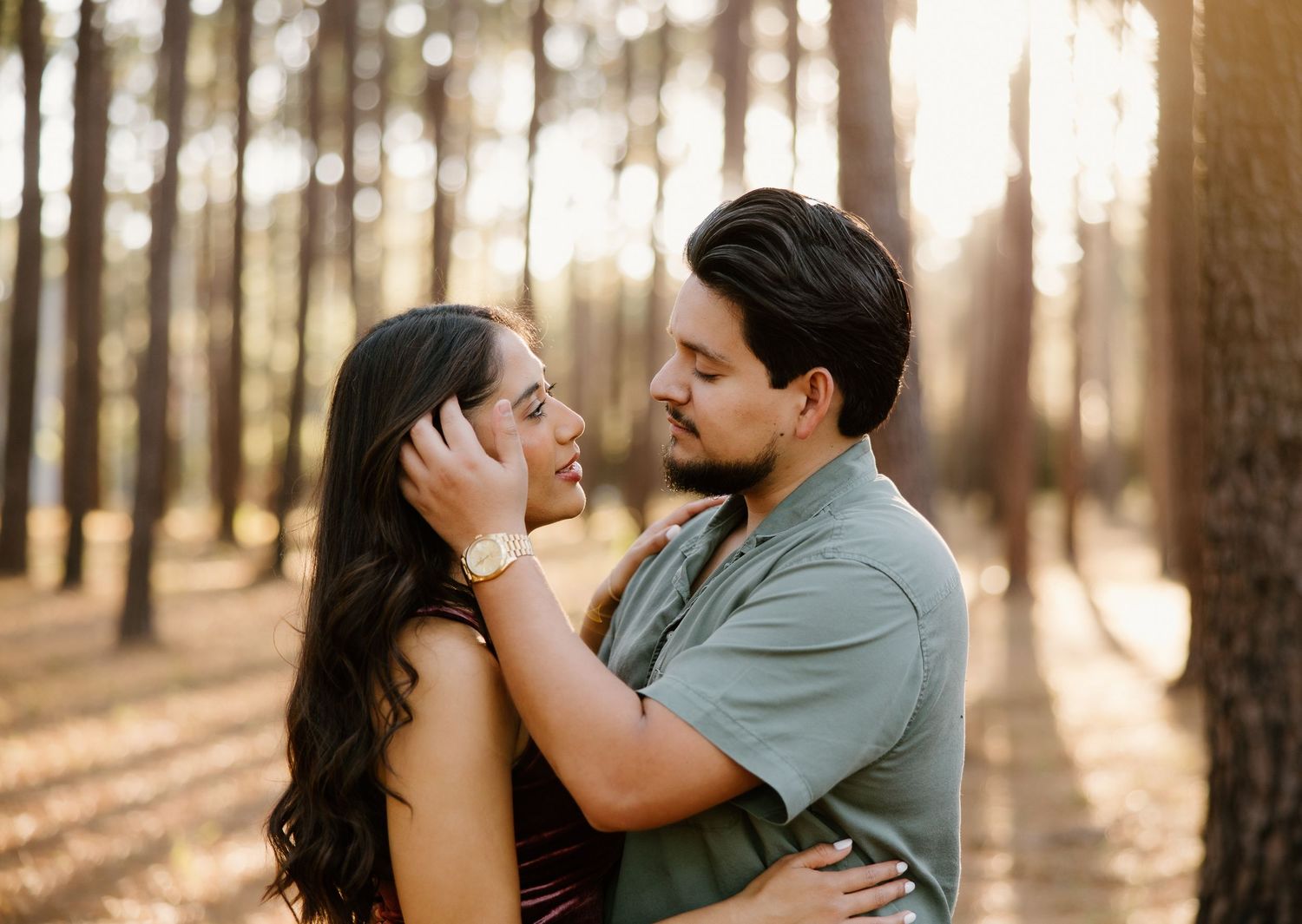 Couple embracing in a sunlit pine forest, woman gently touching man's face.
