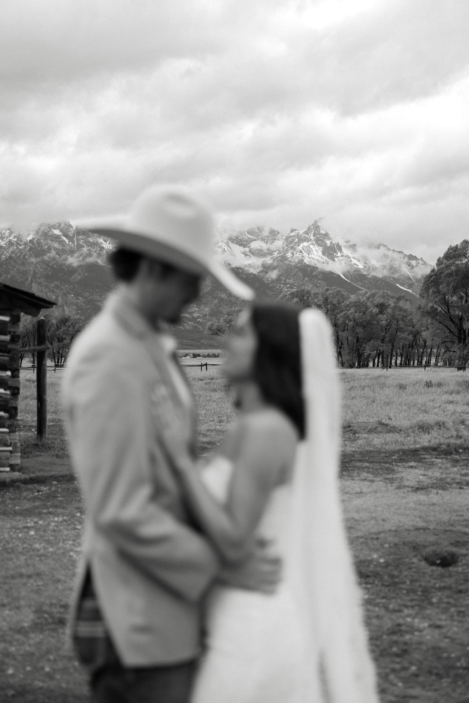 Black and white photo of a couple embracing in cowboy hats with mountain scenery behind them.