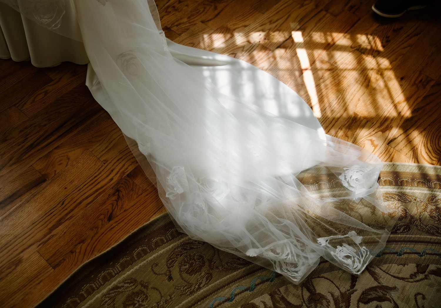 Wedding dress train and veil spread across a wooden floor in warm sunlight.