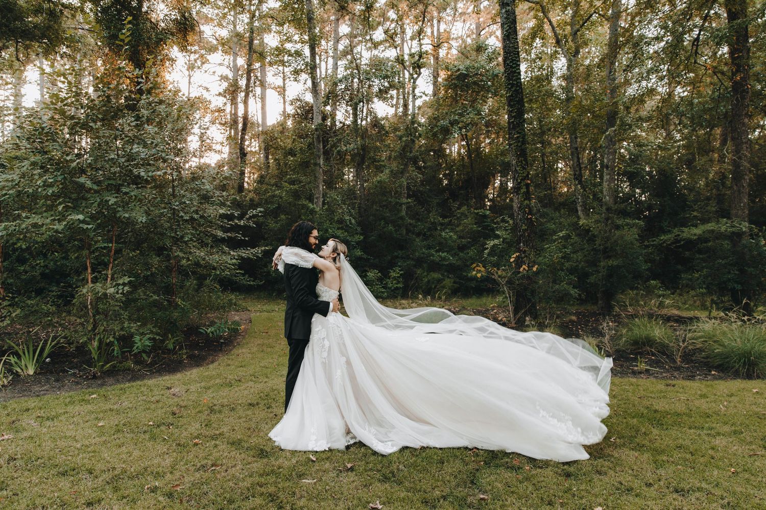 Bride and groom share a romantic embrace outdoors, with the wedding veil flowing dramatically in a lush green forest.