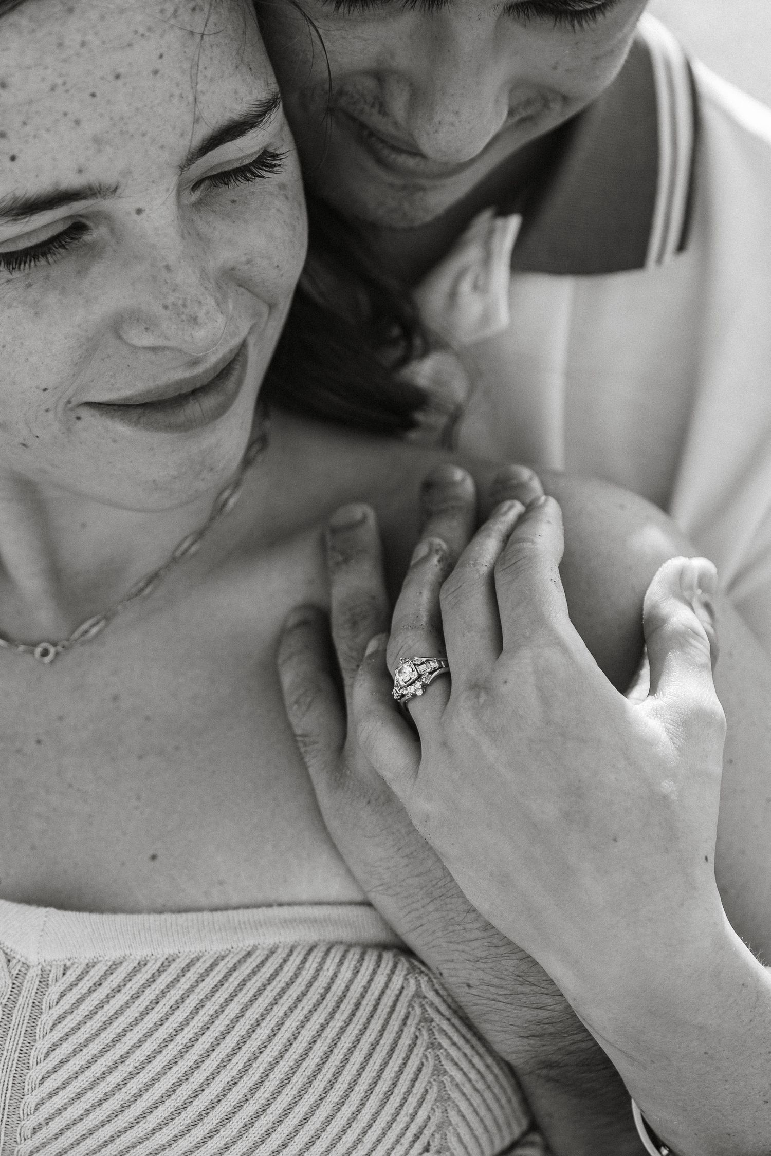 Black and white close-up of couple embracing, woman showing engagement ring on her hand.