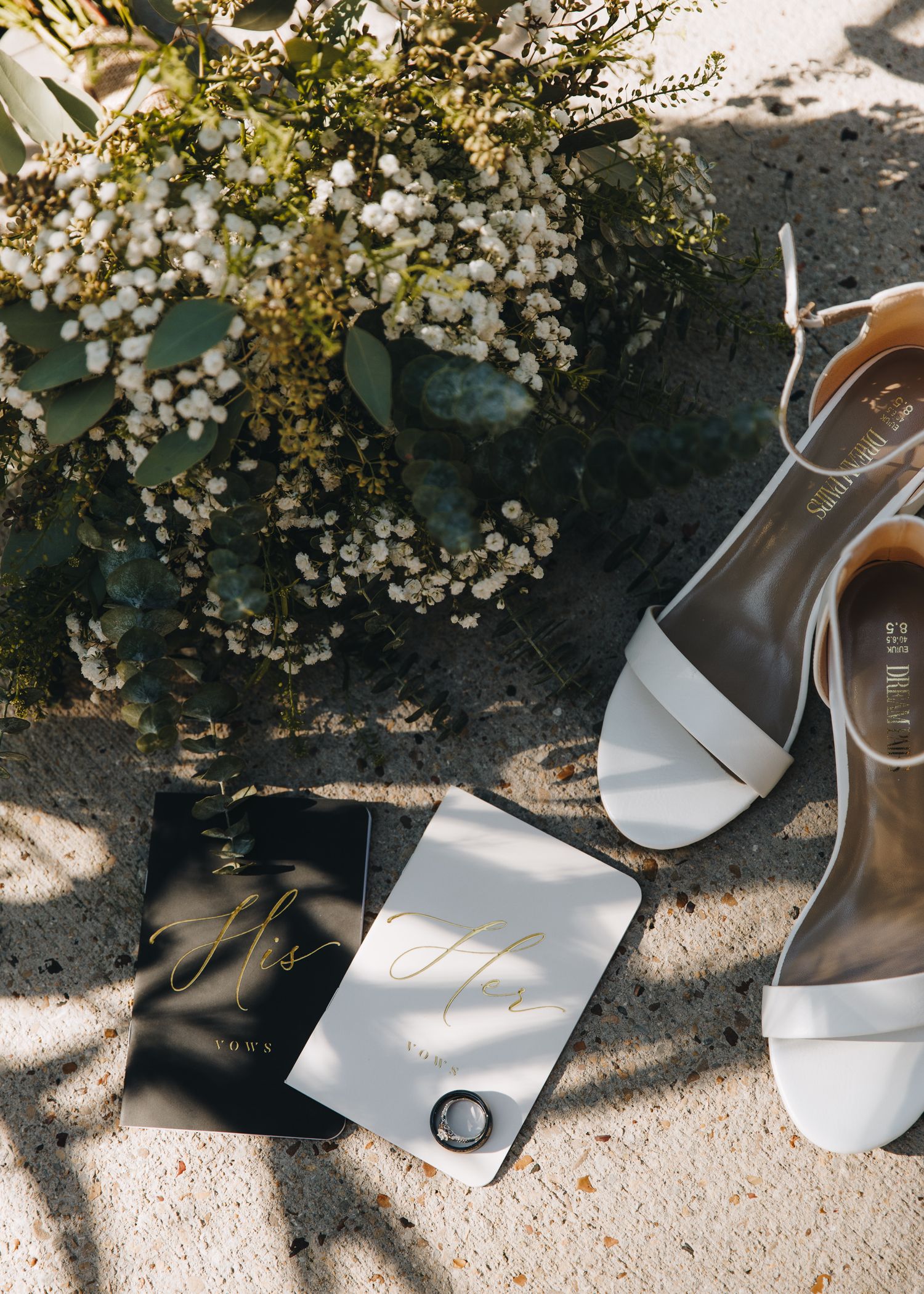 Flat lay of white bridal heels, wedding stationery with gold calligraphy, ring, and floral bouquet.