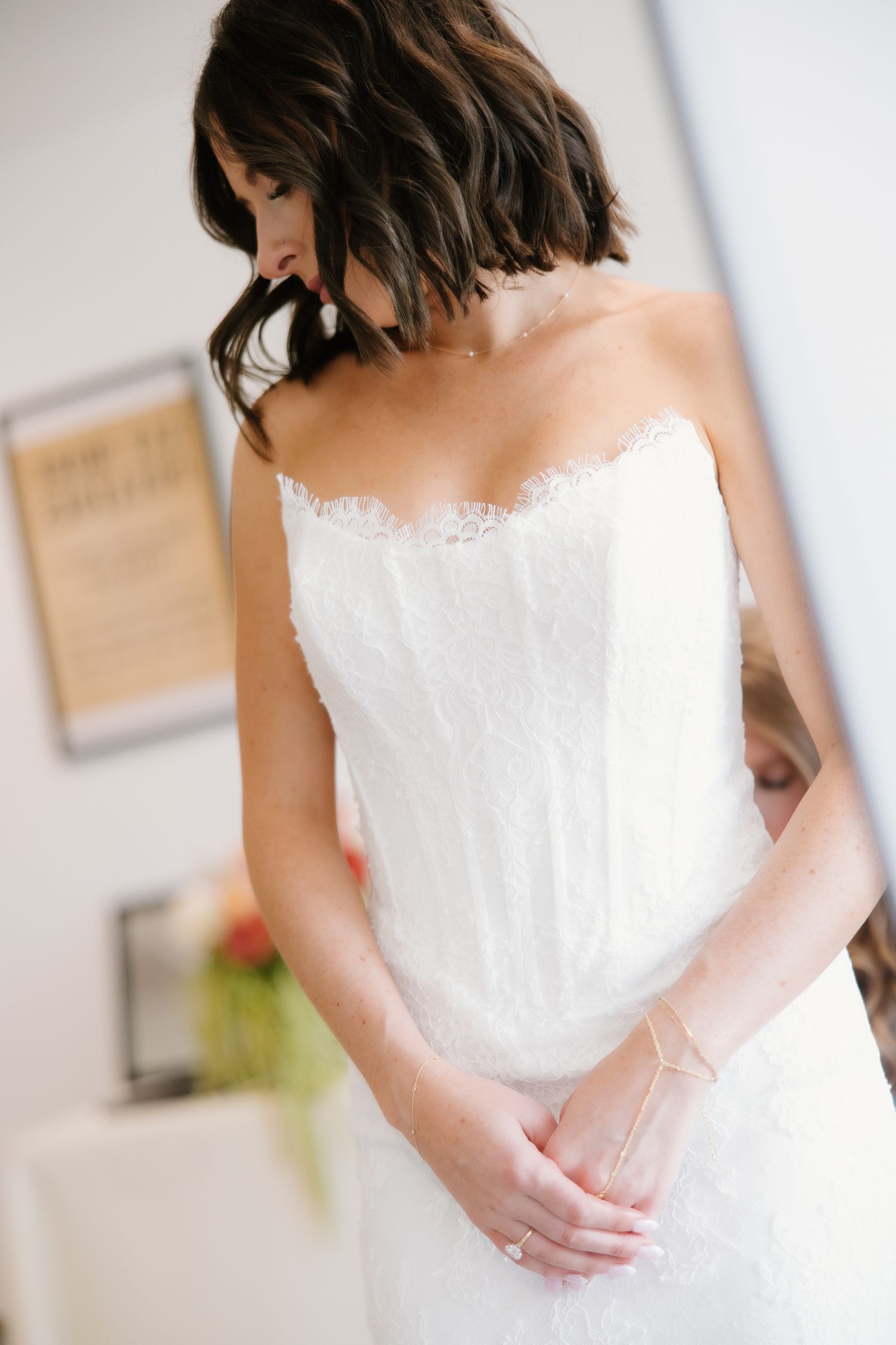 Bride in strapless lace wedding gown looking down with hands clasped, showcasing elegant bridal style.