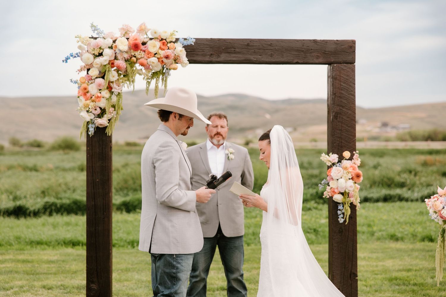 Bride and groom exchanging vows under a rustic wood floral arch outdoors with rolling green hills backdrop.