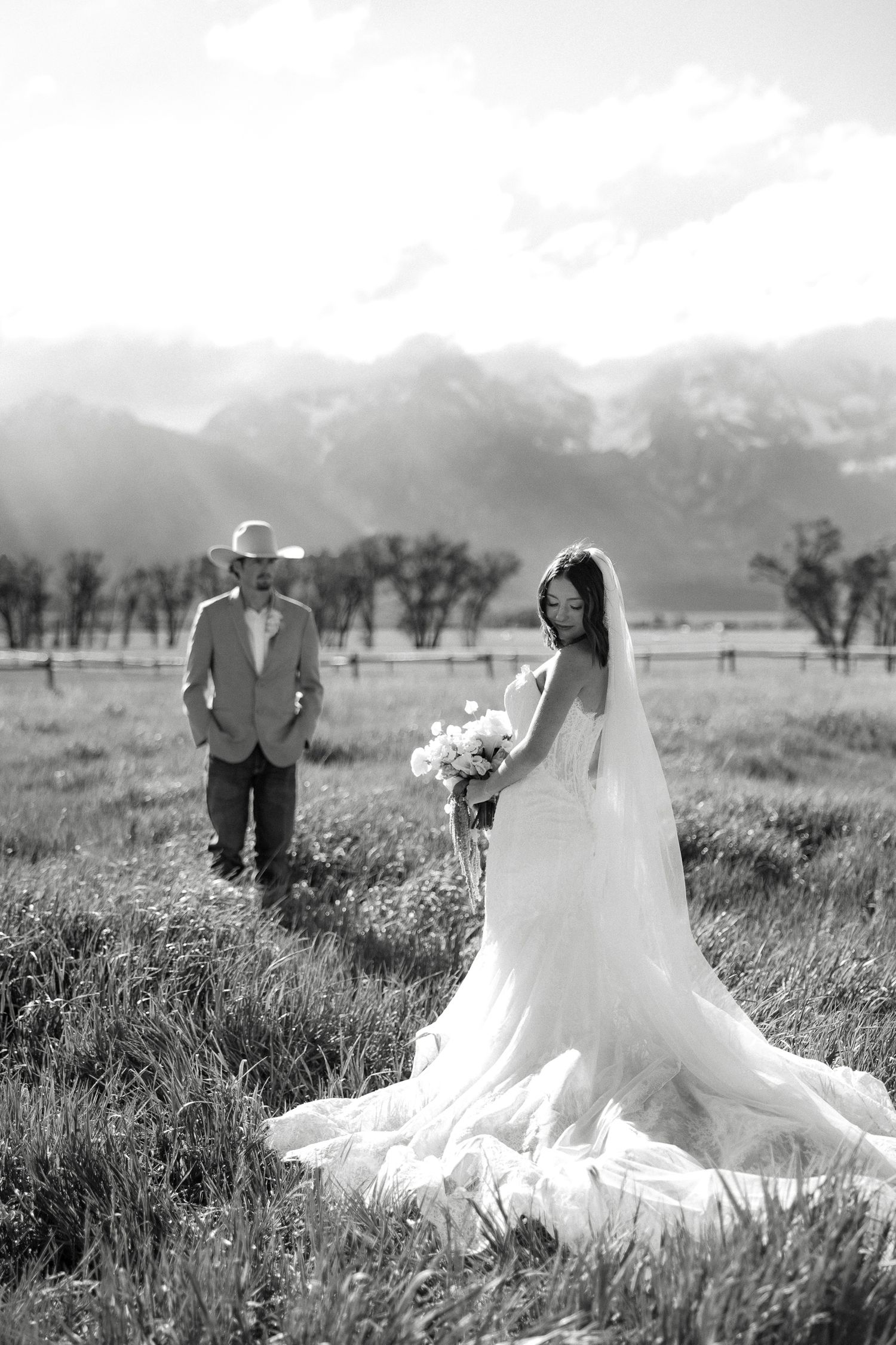 Black and white photo of bride in flowing gown and groom in cowboy hat in mountain meadow.