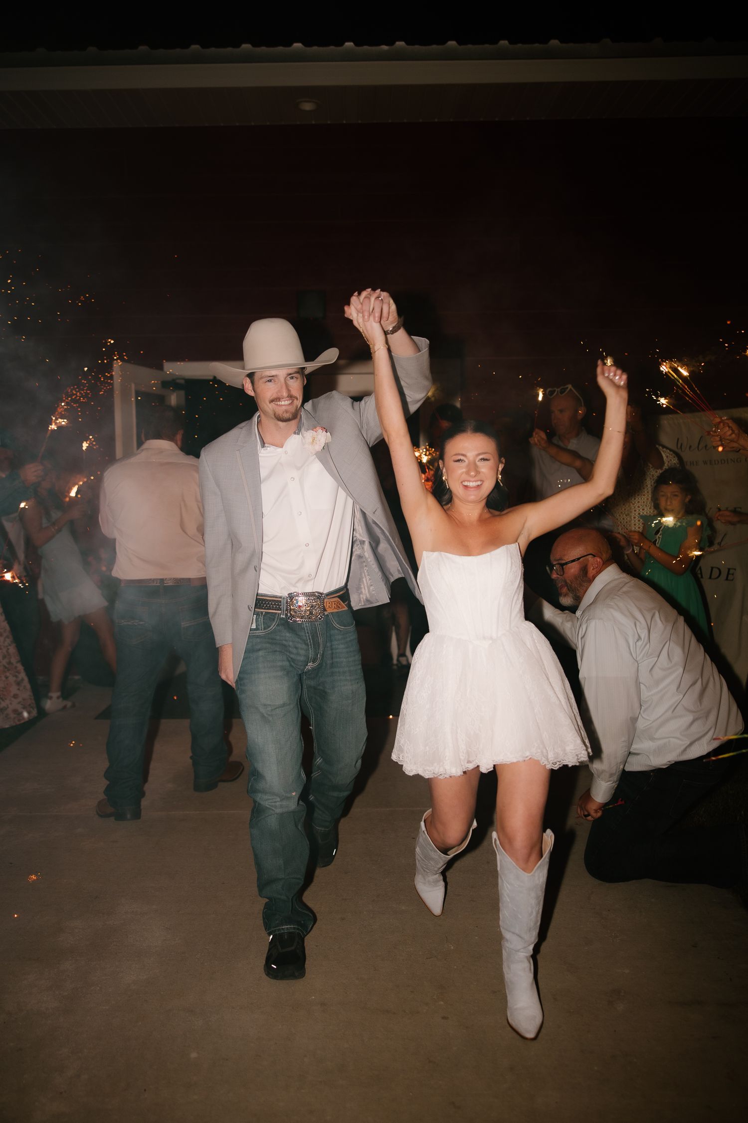 Bride in white mini dress and cowboy boots celebrating with groom in cowboy hat amid sparklers at night.