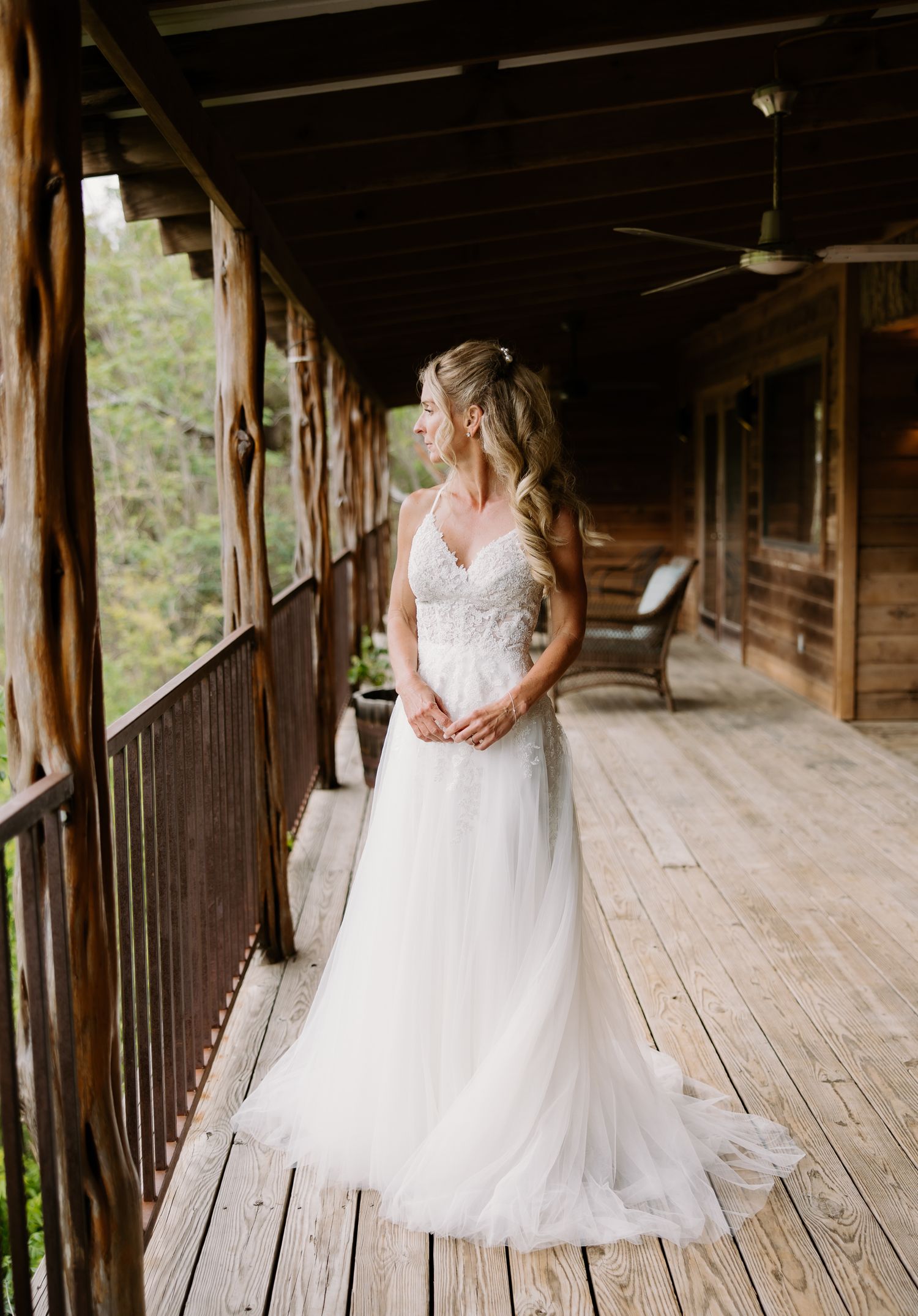 Bride in a lace A-line wedding gown standing on a rustic wooden cabin porch surrounded by lush greenery.