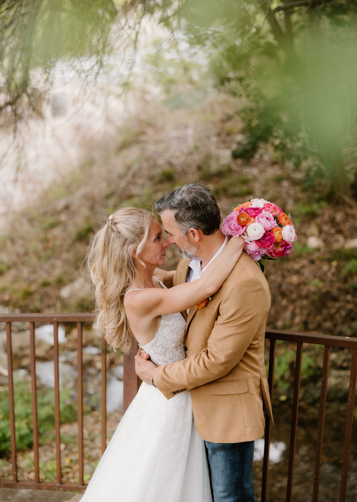 Bride and groom embracing on bridge, groom in tan blazer, bride holding colorful bouquet.