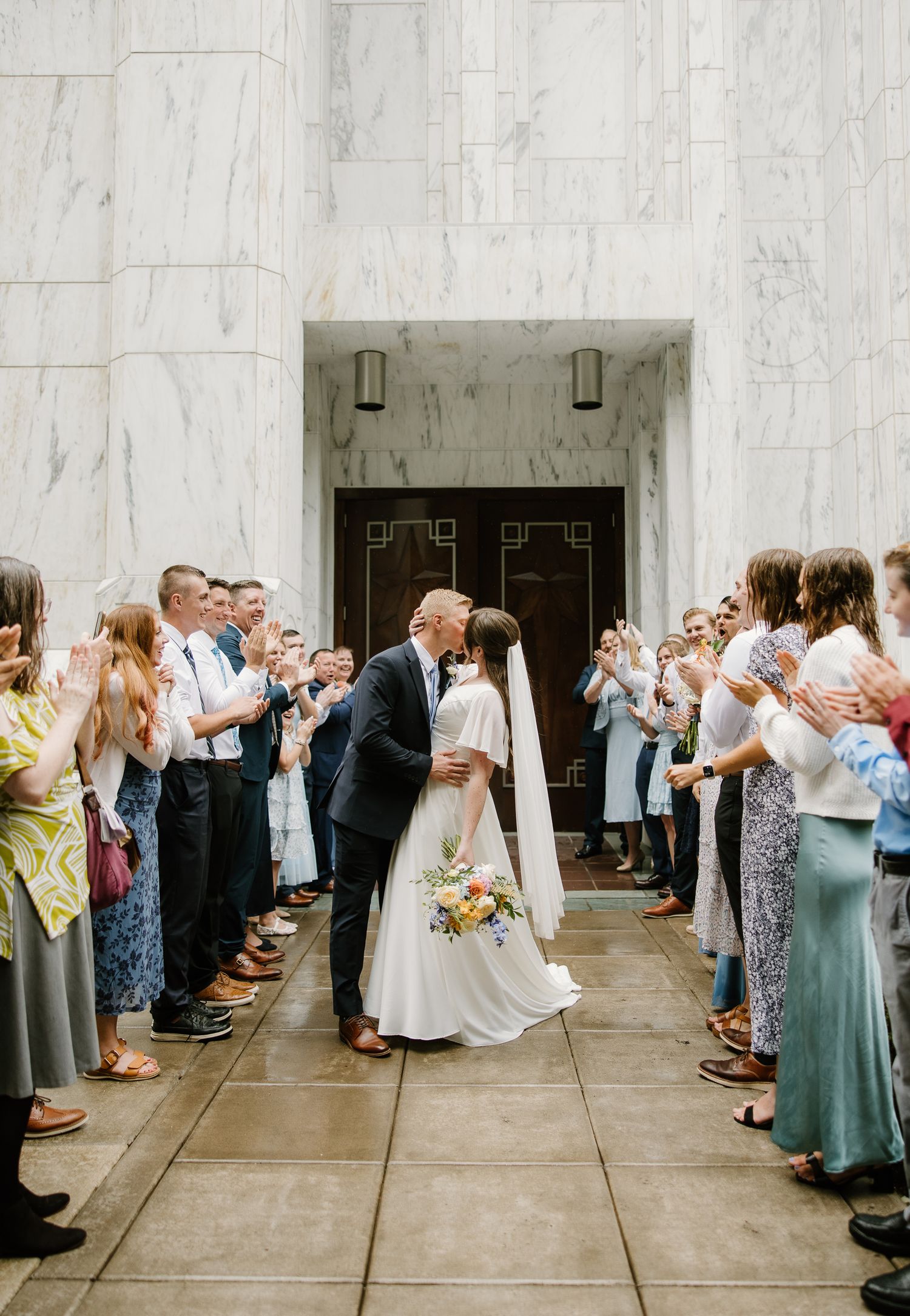 Newlywed couple sharing a kiss outside a marble building while guests applaud during their wedding exit ceremony.