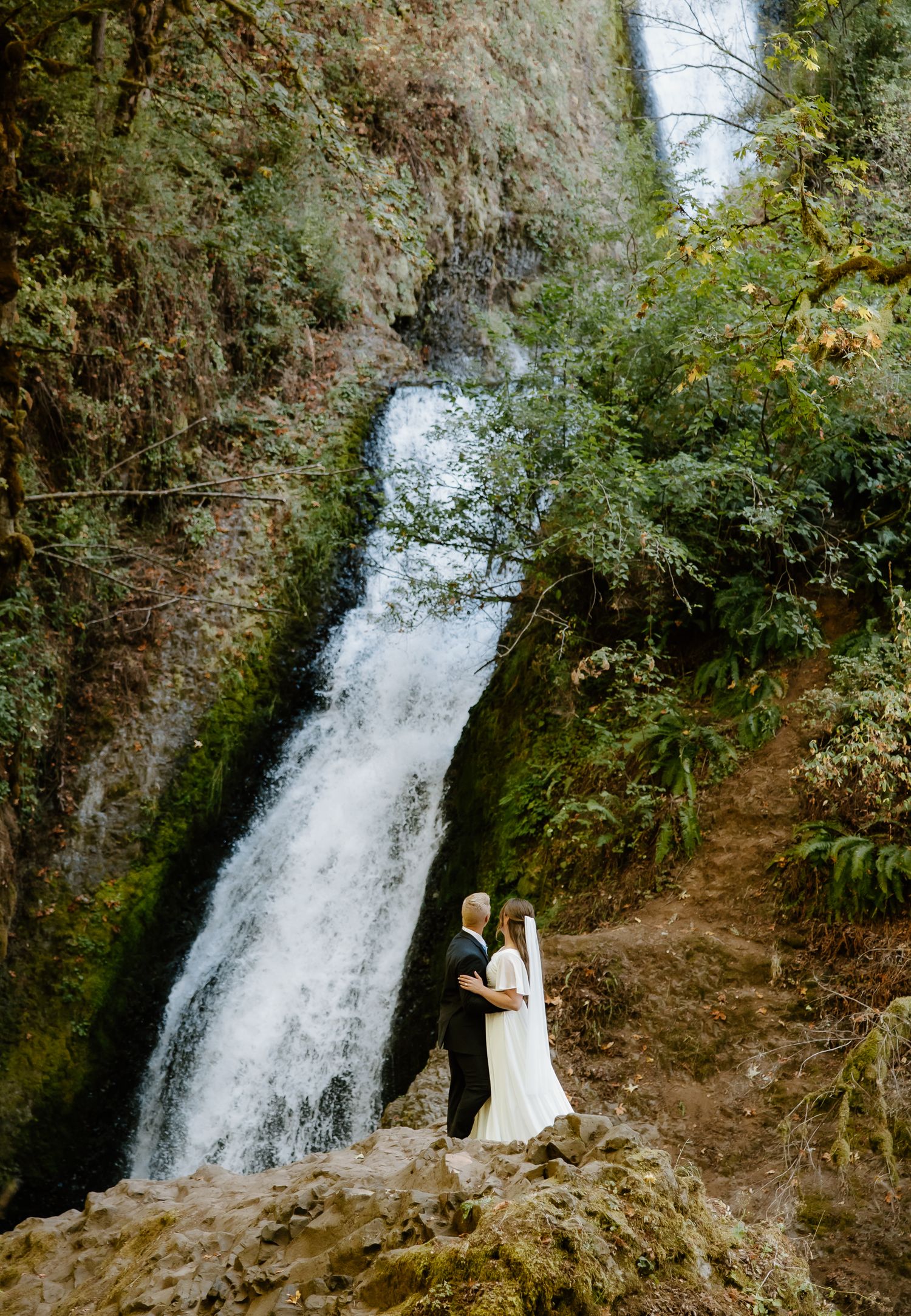 Wedding couple embracing in front of a stunning waterfall surrounded by lush green forest scenery.