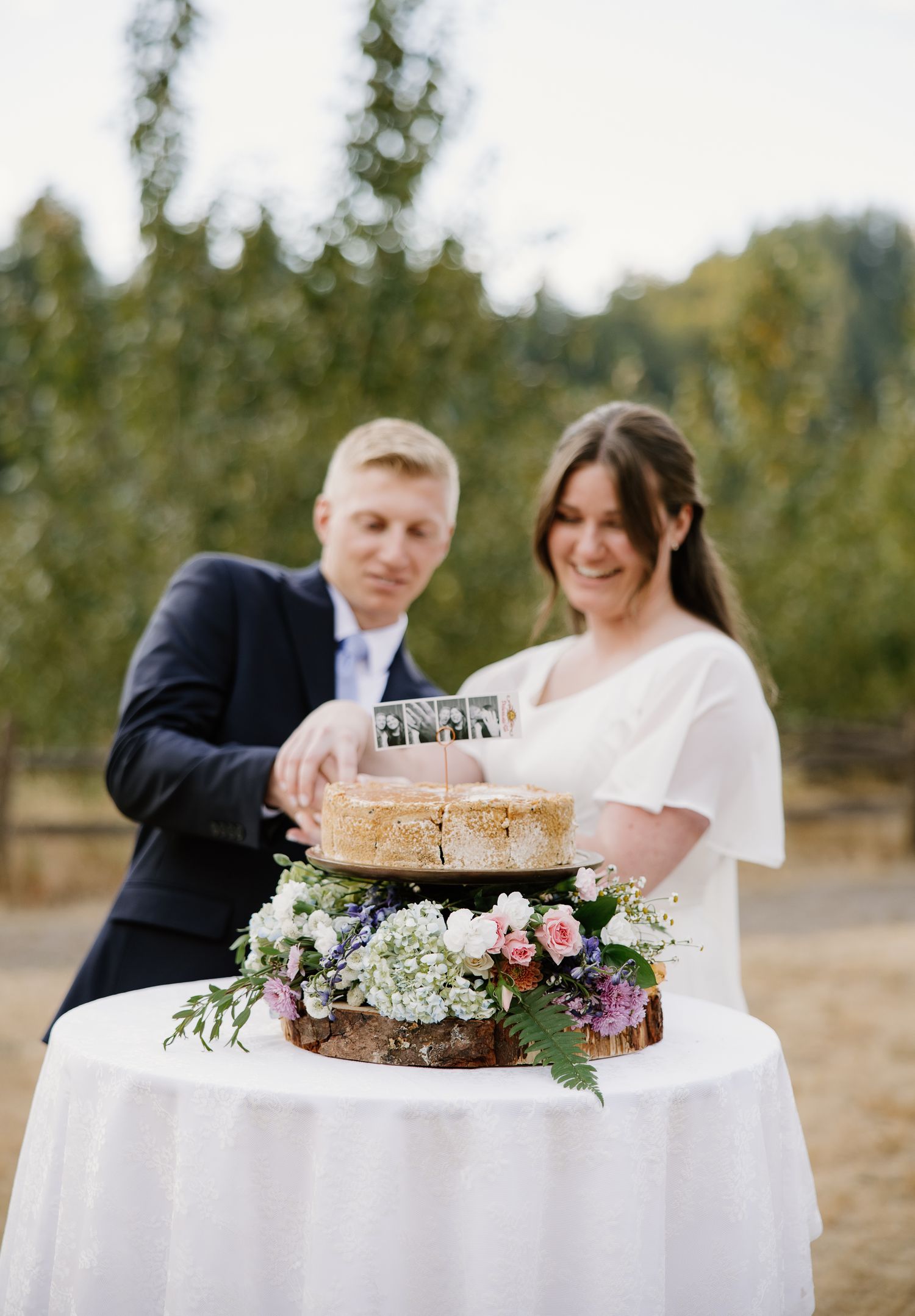 Bride and groom cut rustic naked wedding cake decorated with flowers on a wooden slice stand outdoors.
