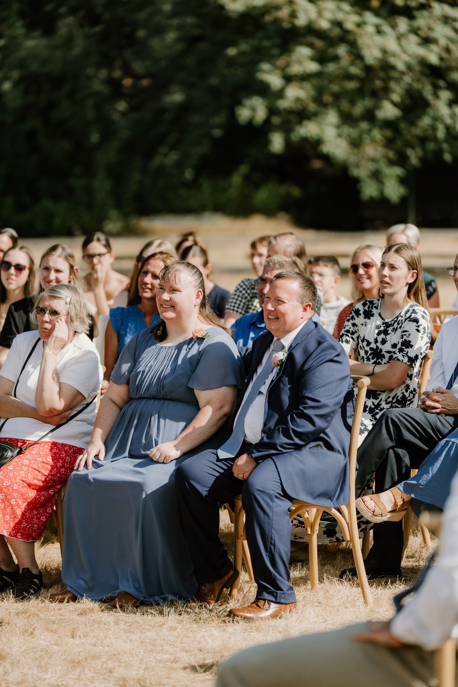 Smiling wedding guests in blue attire sit together during an outdoor wedding ceremony.