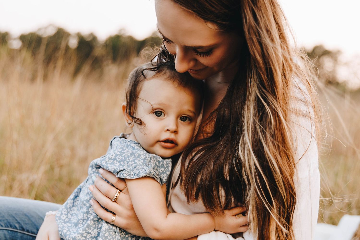Mother tenderly embraces her curly-haired toddler daughter in a warm golden field portrait.