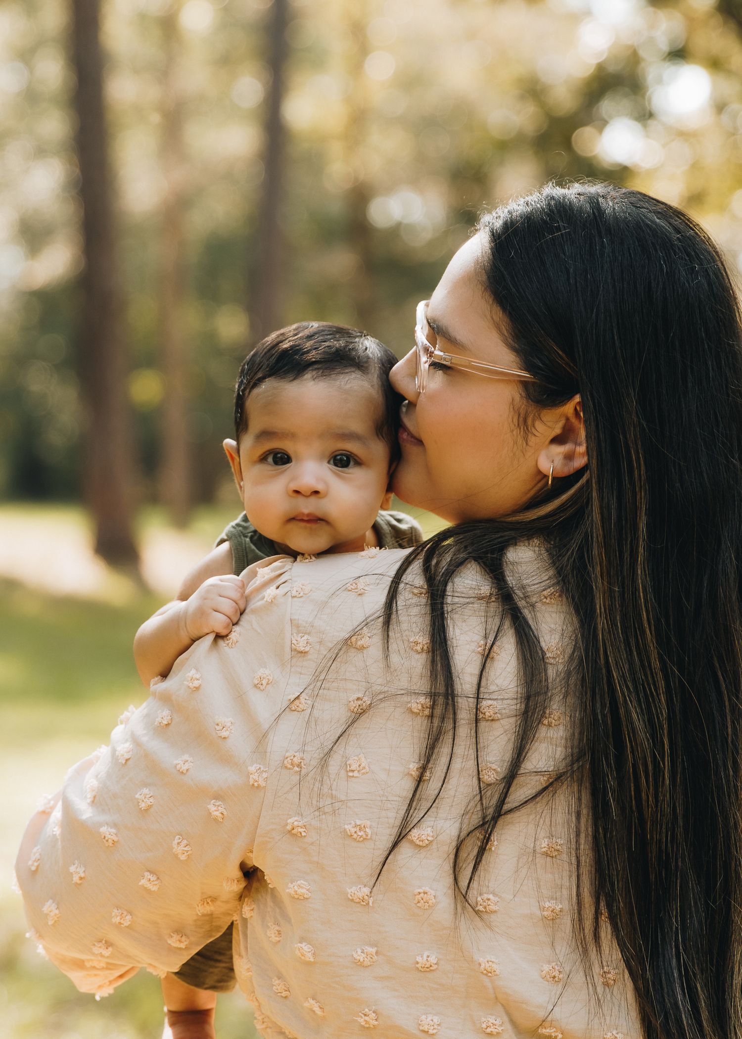 Mother with long dark hair and glasses kisses her baby in a sunlit outdoor portrait.
