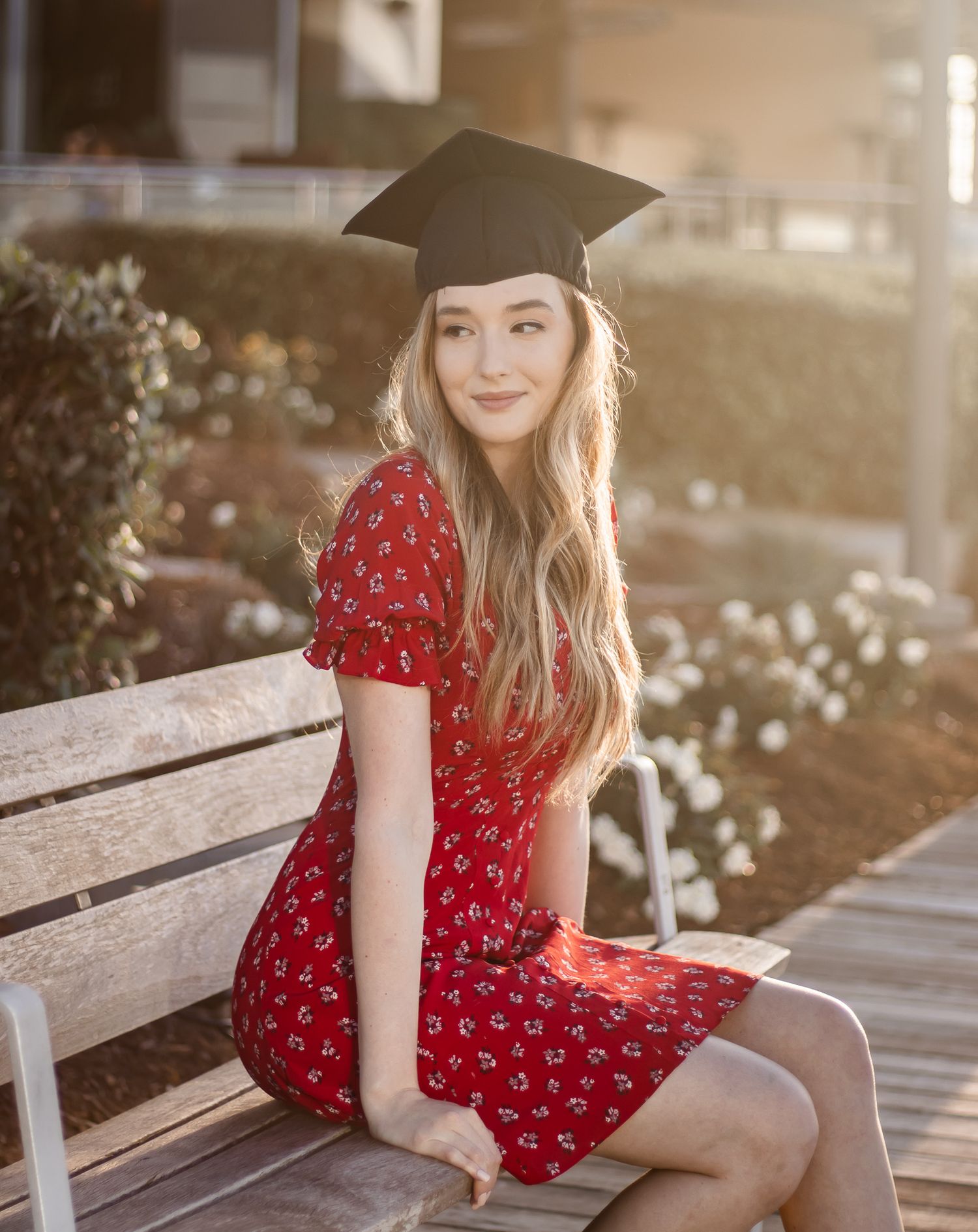 Smiling graduate in red floral dress and cap sits on bench in golden sunlight.