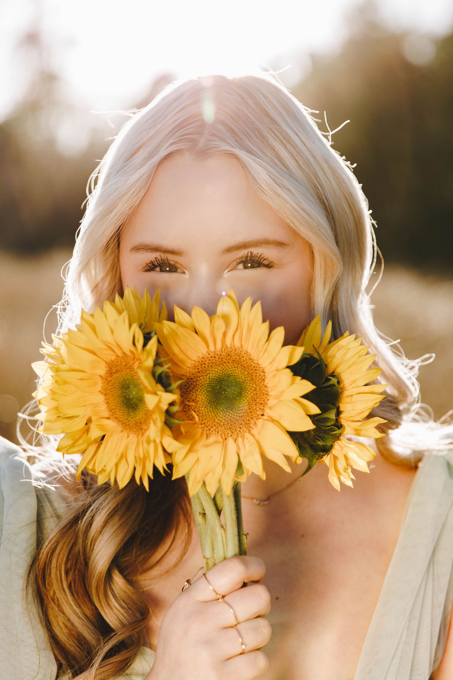 Blonde woman holding bright yellow sunflowers in front of her face in golden sunlight.