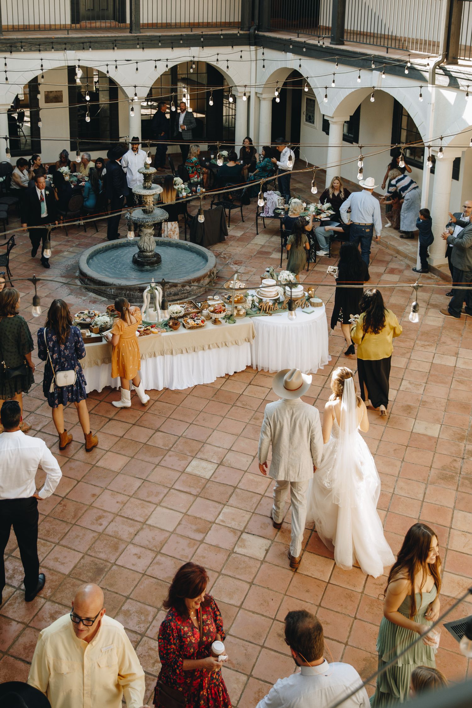 Aerial view of wedding reception in a courtyard with fountain, string lights, buffet table, and guests mingling.