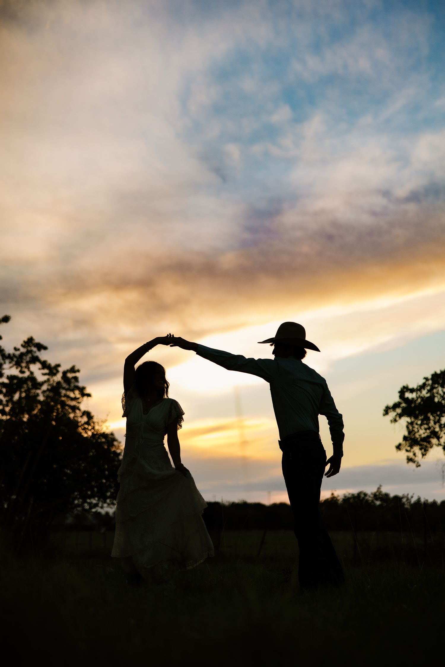 Silhouette of couple dancing at sunset, man in cowboy hat holding woman's hand.