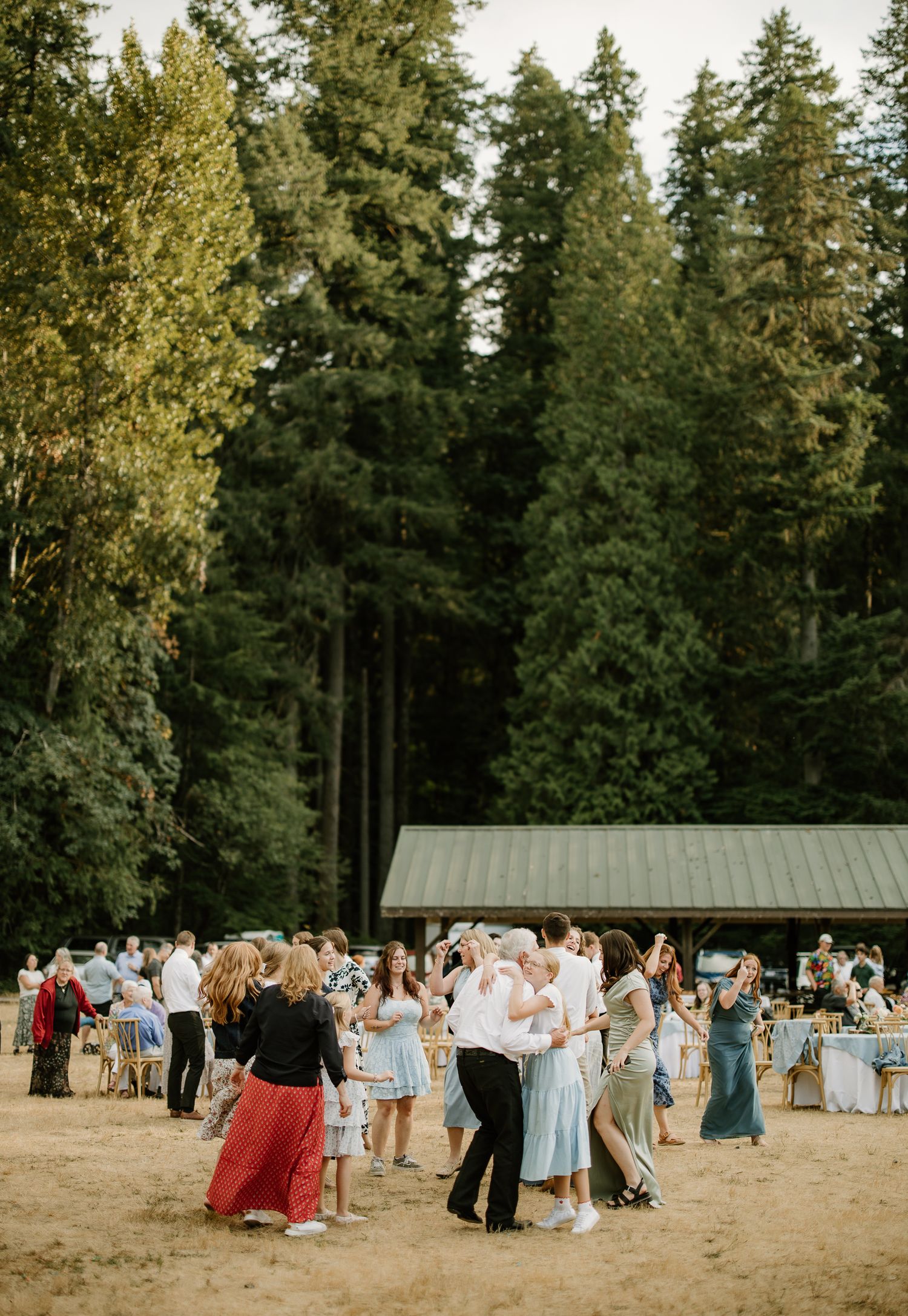 Wedding guests dance outdoors on sandy ground surrounded by tall evergreen trees near a rustic pavilion.