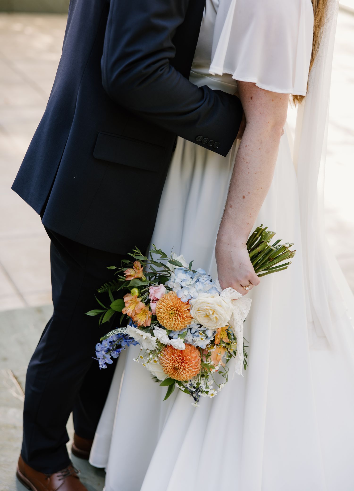 Bride and groom embrace closely as bride holds a colorful bouquet of dahlias and hydrangeas.