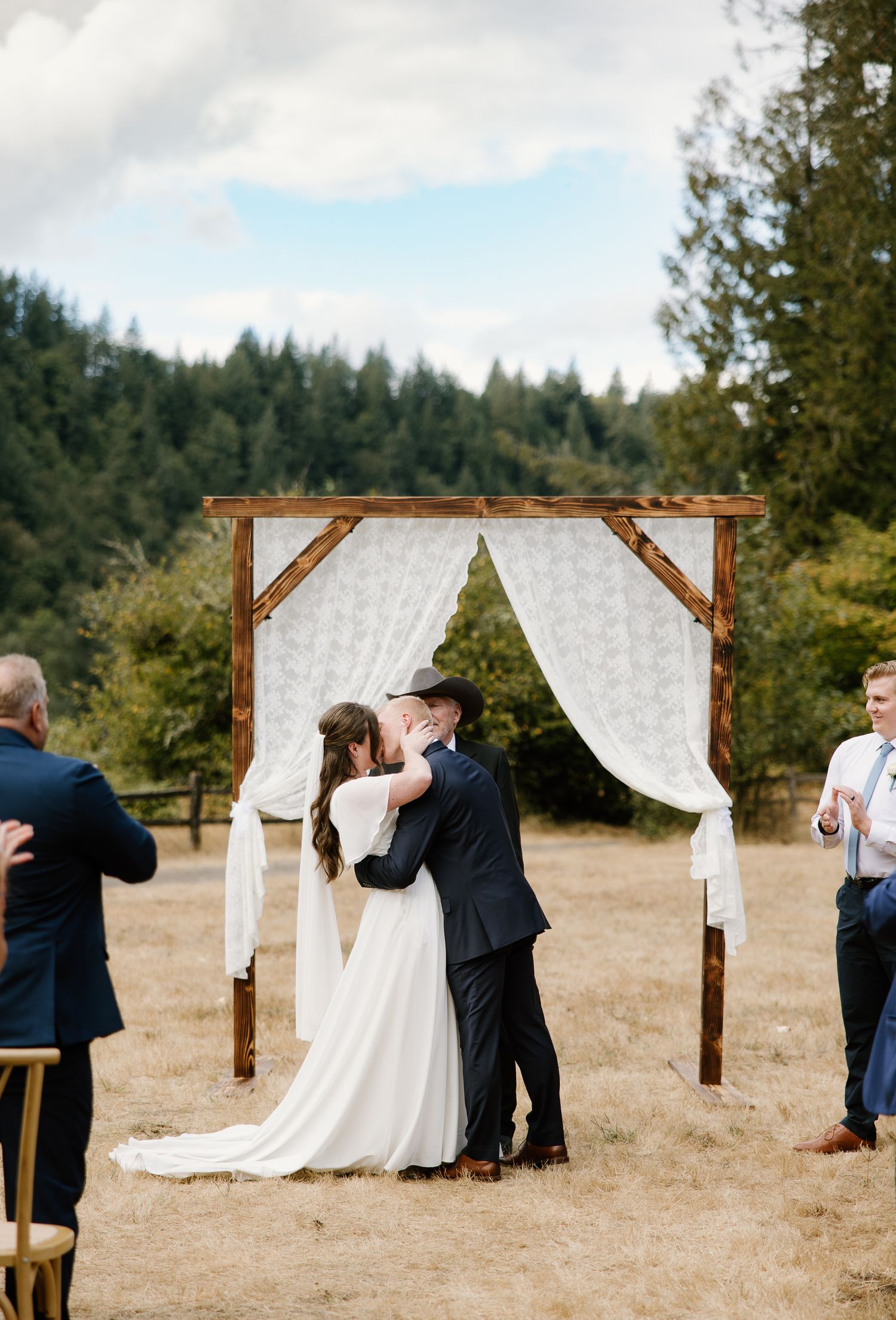 Newlyweds share first kiss under rustic wooden arch with white draping at outdoor forest wedding.