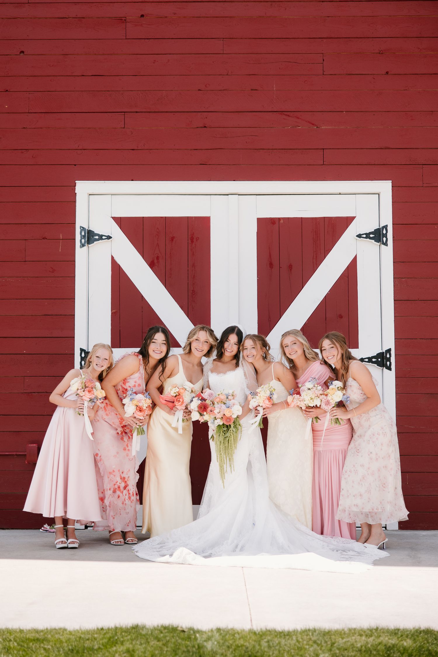 Bride and bridesmaids in mismatched pink dresses holding floral bouquets in front of a red barn.