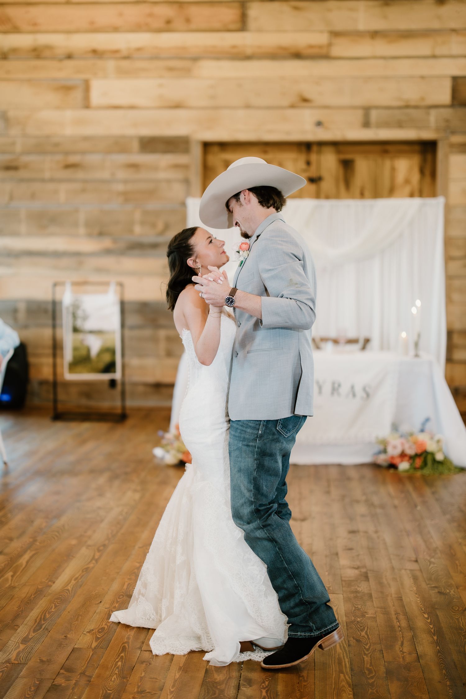 Bride and groom share their first dance in a rustic wooden barn reception venue with warm lighting.
