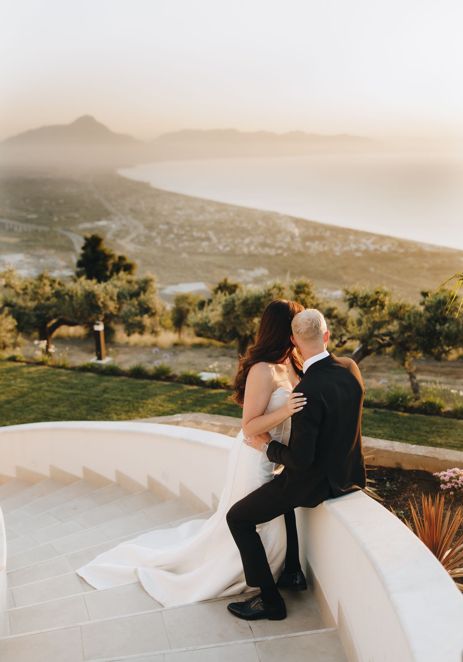 Bride and groom sit together on a terrace overlooking a stunning coastal mountain sunset view.