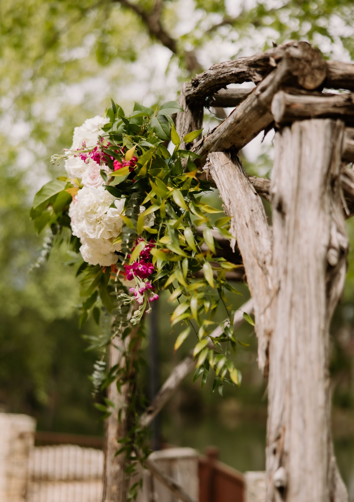 Rustic driftwood wedding arch adorned with white hydrangeas, pink flowers, and lush greenery outdoors.