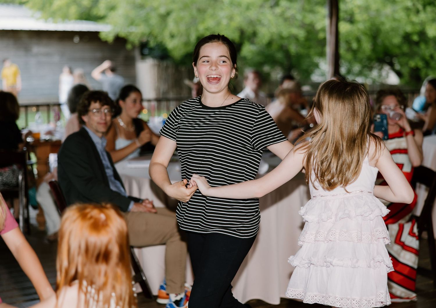 Young girl in a striped shirt dancing joyfully with a child in a white dress at a wedding reception.