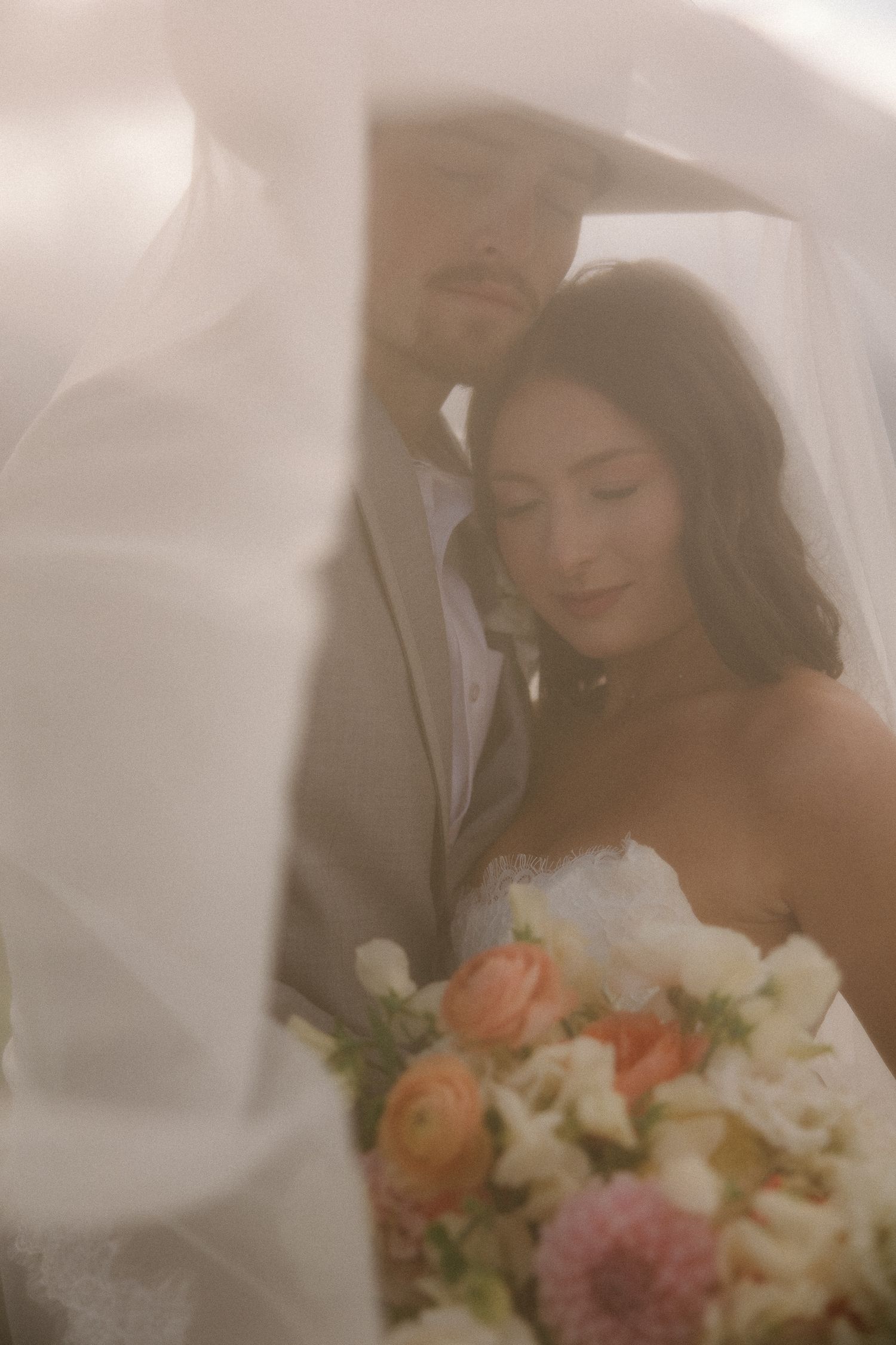 Bride and groom share an intimate moment under a flowing veil, bride holding a pastel bouquet of roses and flowers.