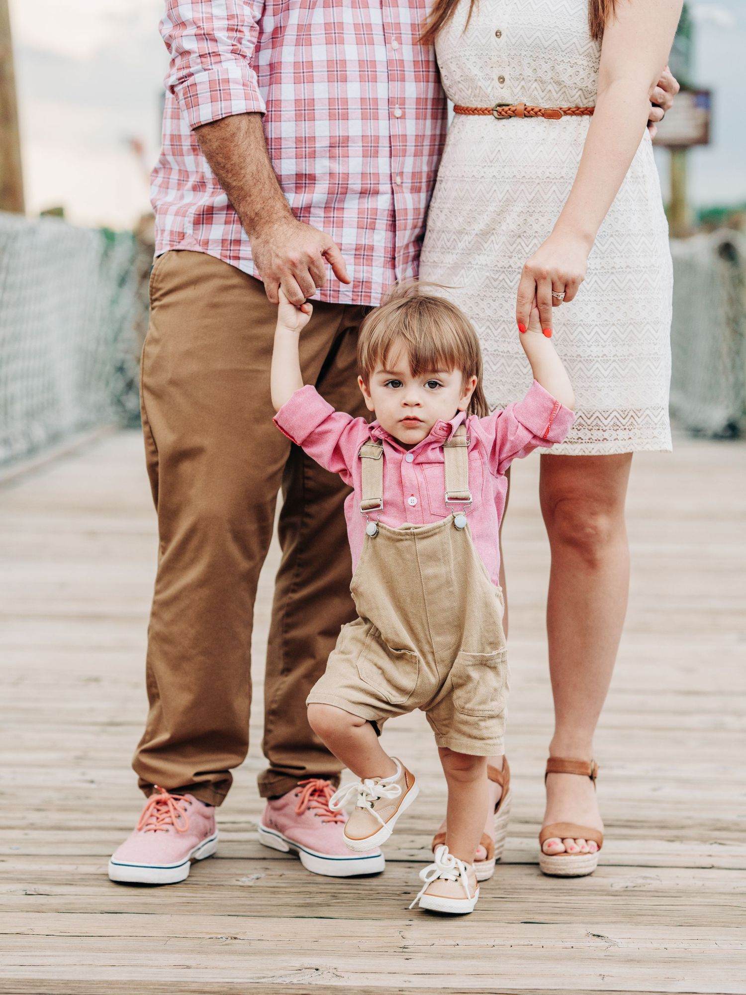 Small child in overalls holds hands with adults while walking on a wooden boardwalk on a sunny day.